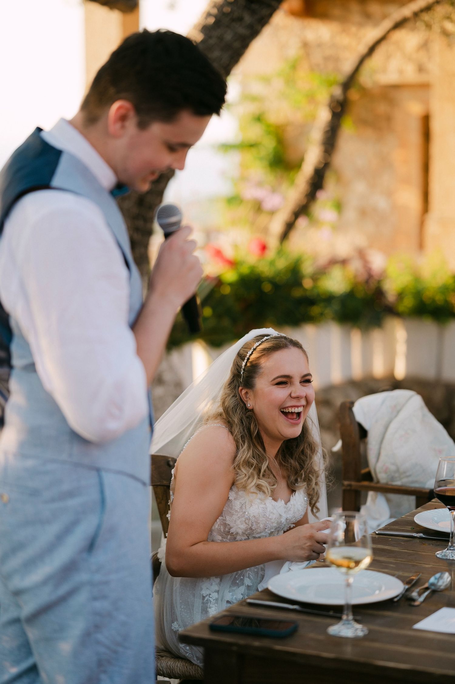 A bride laughs joyfully at an outdoor wedding reception while someone gives a speech at the rustic wooden table.