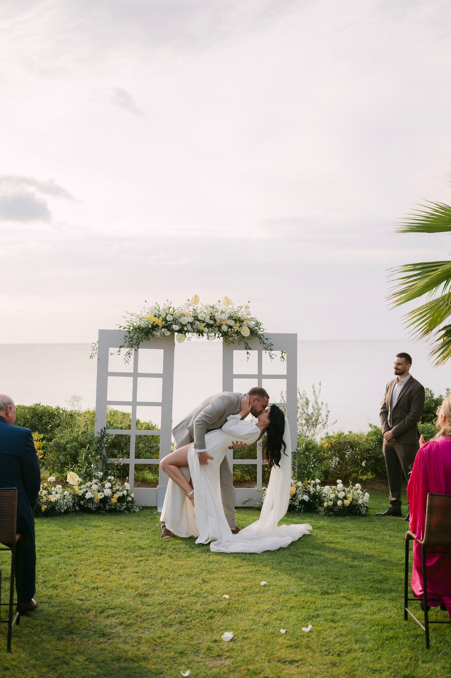 Outdoor wedding ceremony setup with white window frames and floral arrangements on a grassy lawn by the ocean.