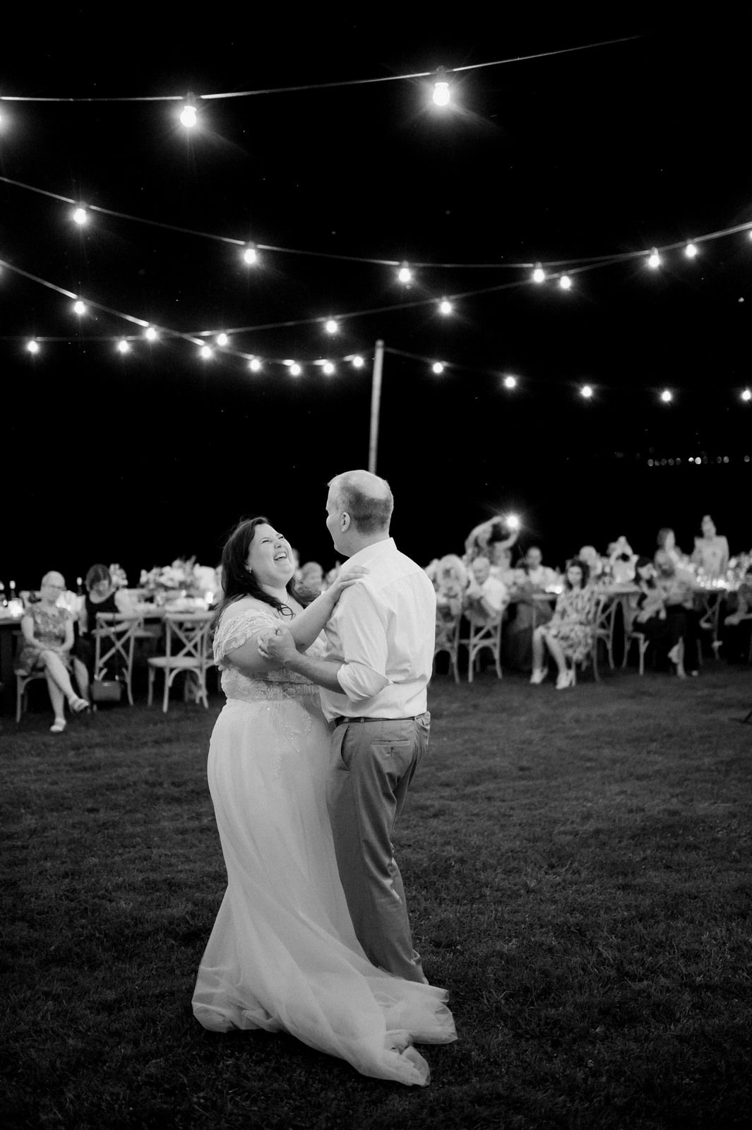 A couple dances under string lights during an outdoor evening reception.
