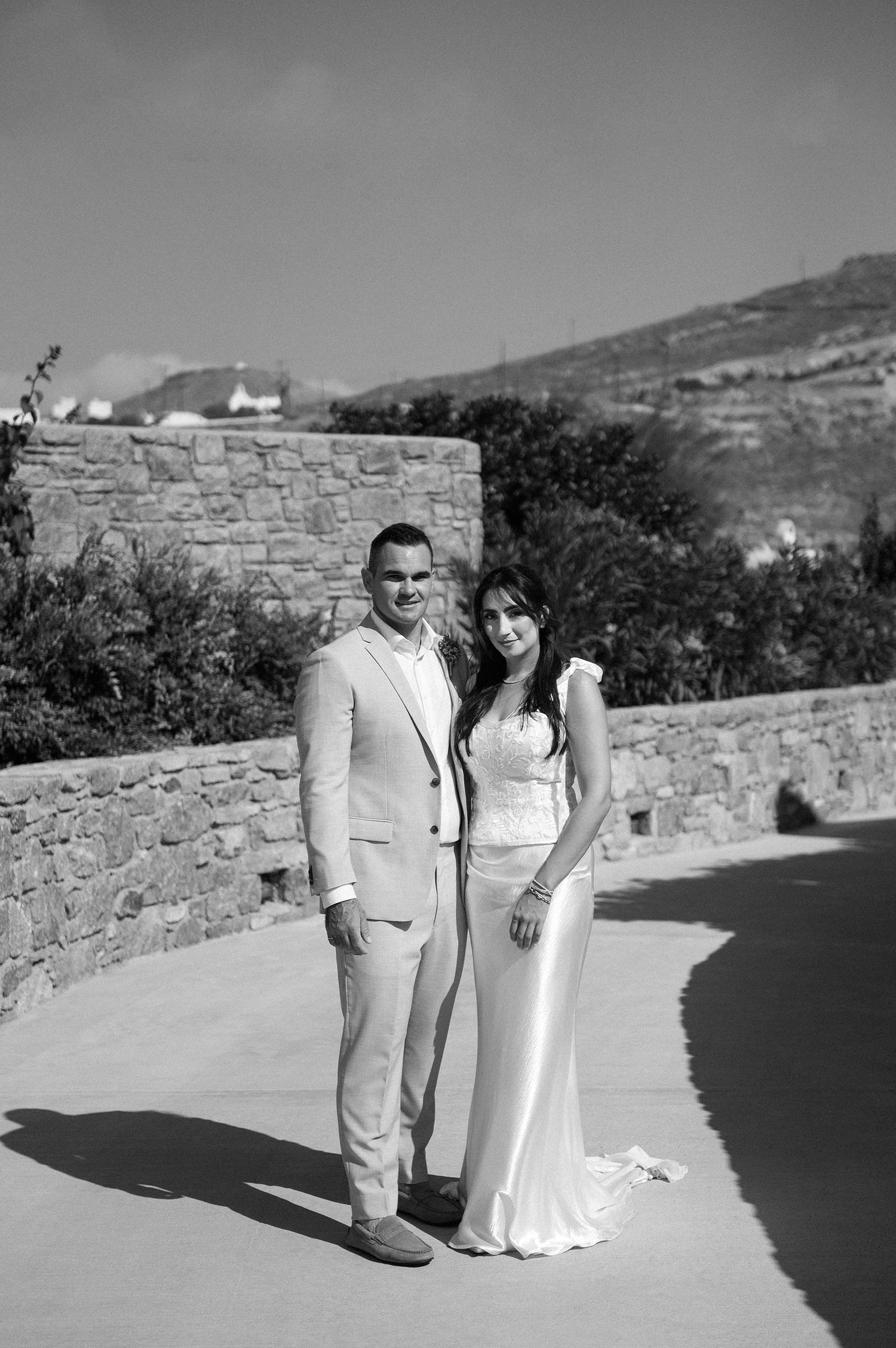 A couple in formal attire poses together against a stone wall backdrop in black and white photography.