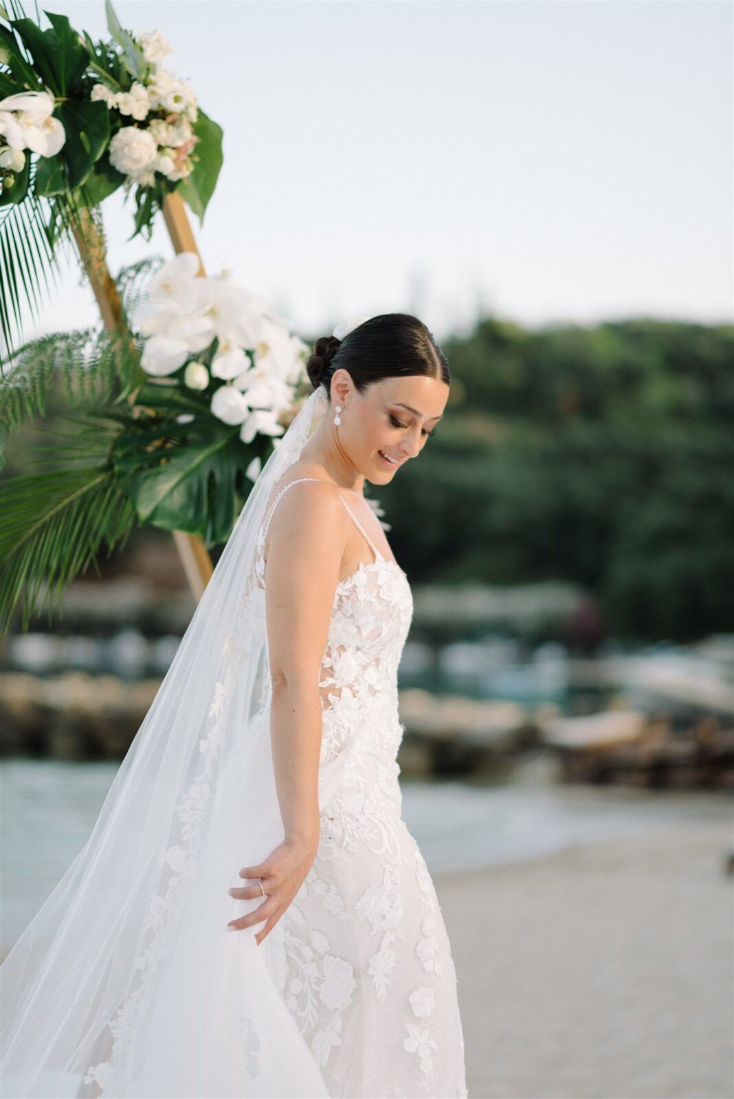 A bride in an elegant lace wedding dress with flowing veil stands gracefully on a sandy beach.