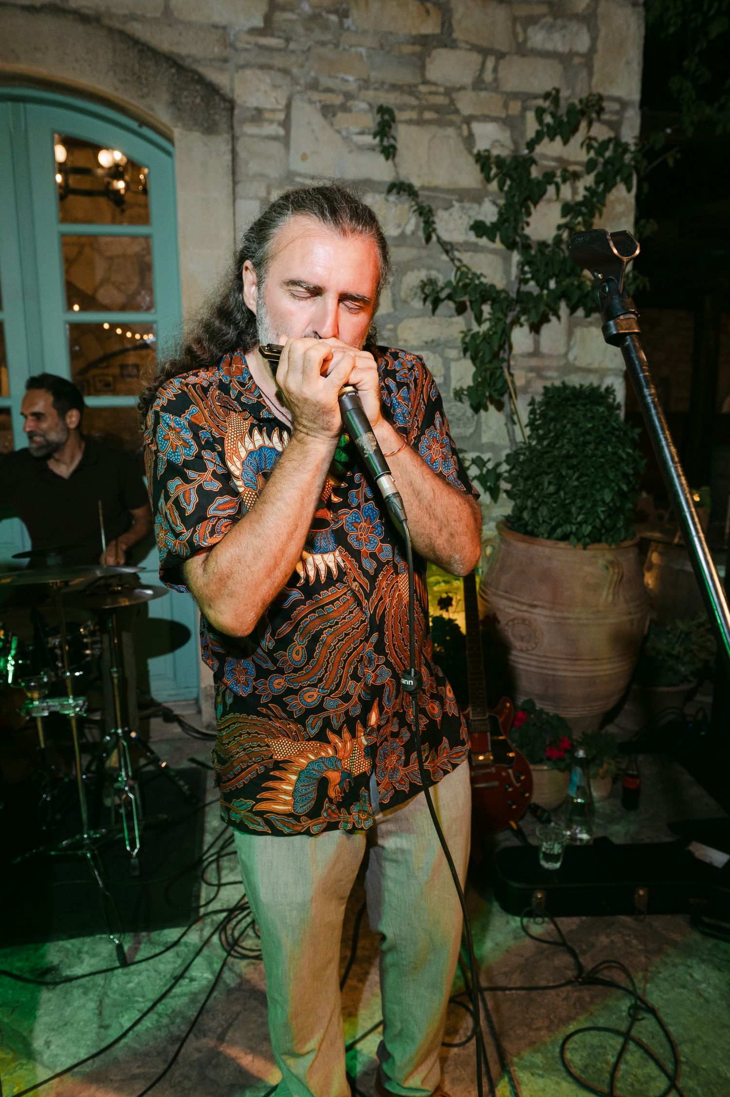 A musician performs on stage playing harmonica at an outdoor venue with stone walls and a turquoise door in the background.