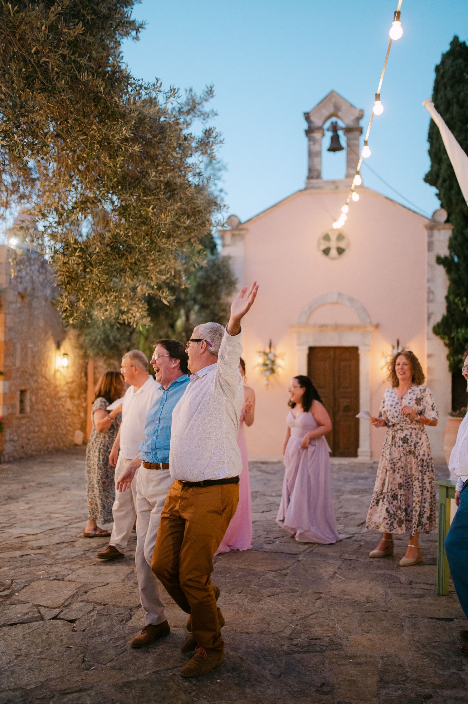 People dancing outside a charming white chapel with string lights at dusk during a celebration.