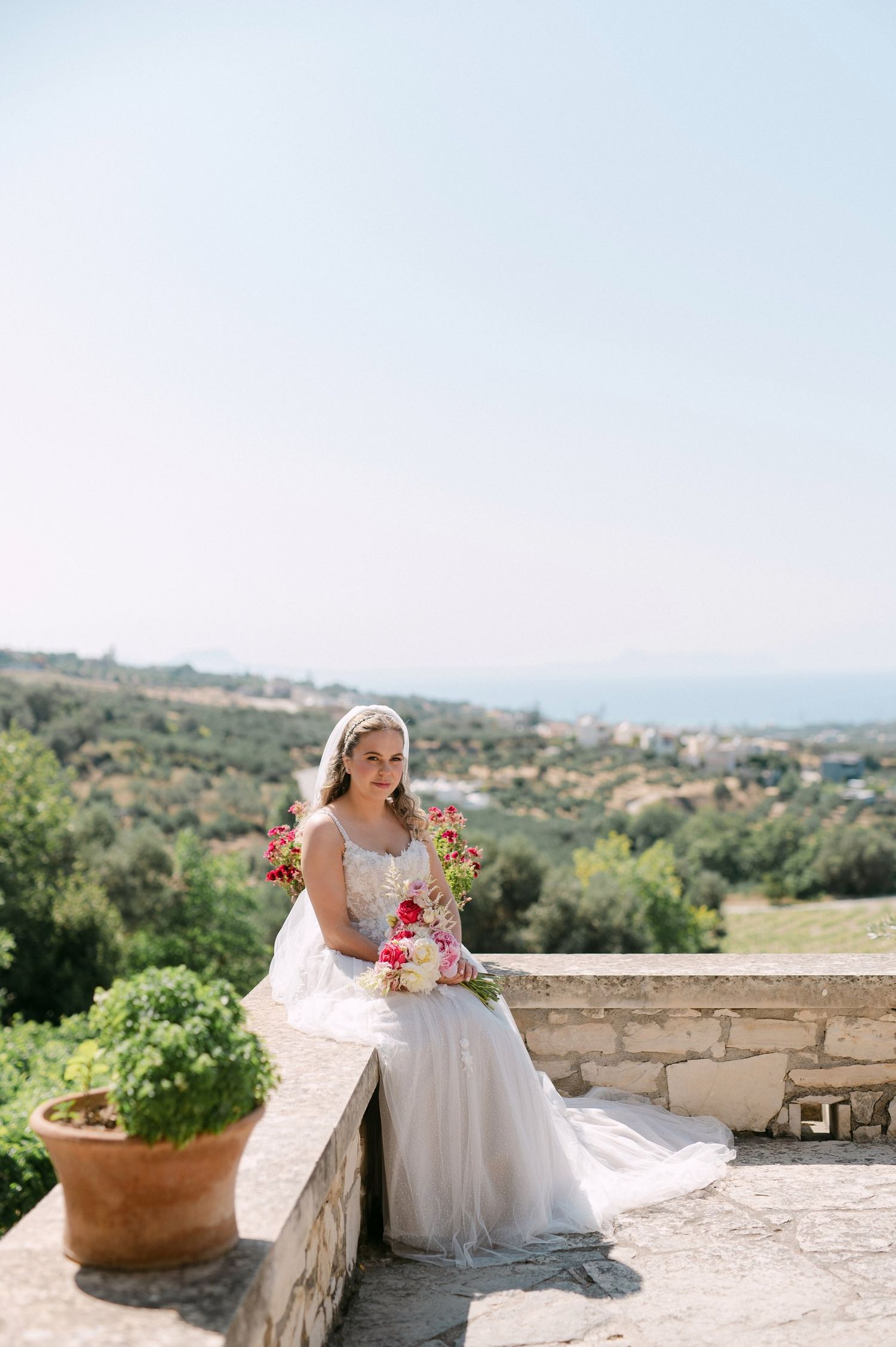 Elegant bride in white gown sits on stone wall at Mediterranean countryside venue holding vibrant bouquet on sunny day.