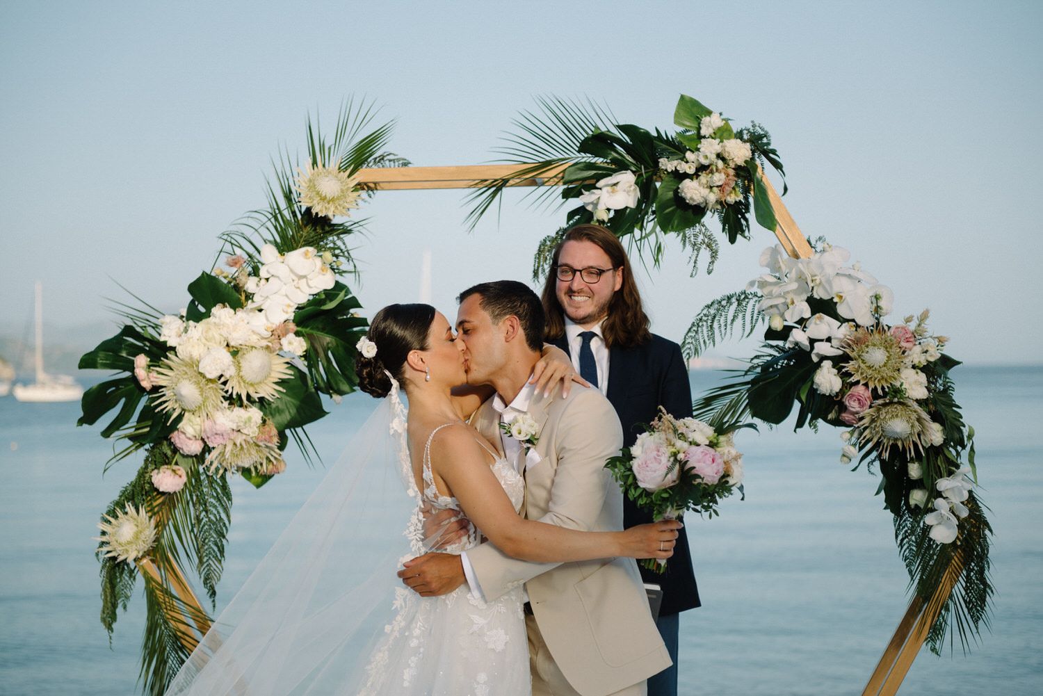 Newlyweds share their first kiss under a floral arch with ocean views and boats in the background.
