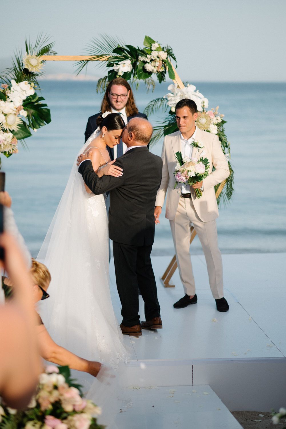 Emotional wedding ceremony moment under floral arch with ocean backdrop.