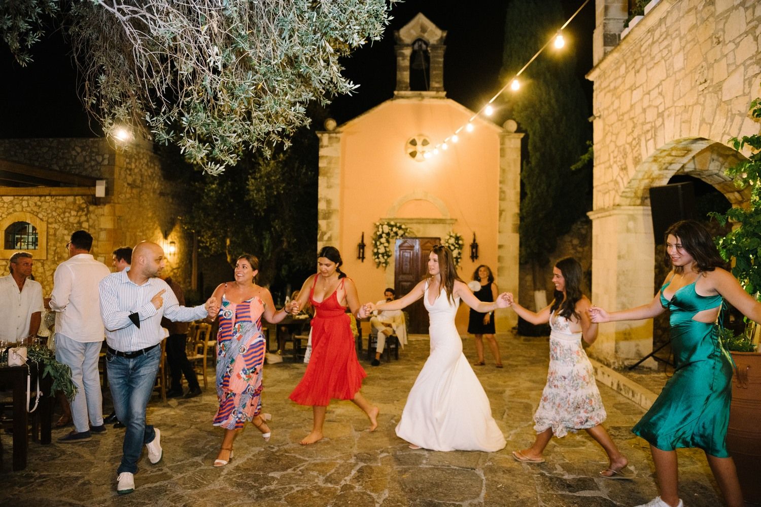 Wedding guests dancing outdoors at night near a small chapel with string lights.
