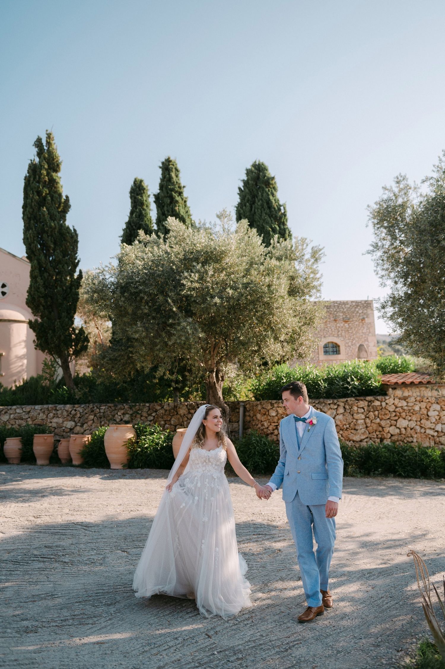 A newlywed couple walks hand in hand through a Mediterranean garden with cypress trees and stone walls on a sunny day.