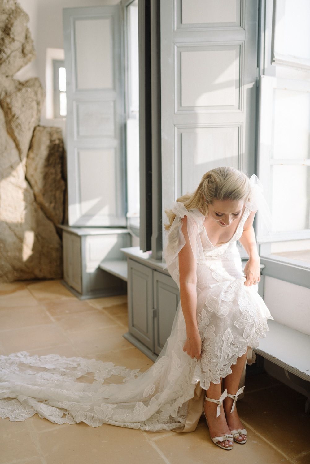Elegant bridal moment adjusting shoes by tall window with trailing dress train.