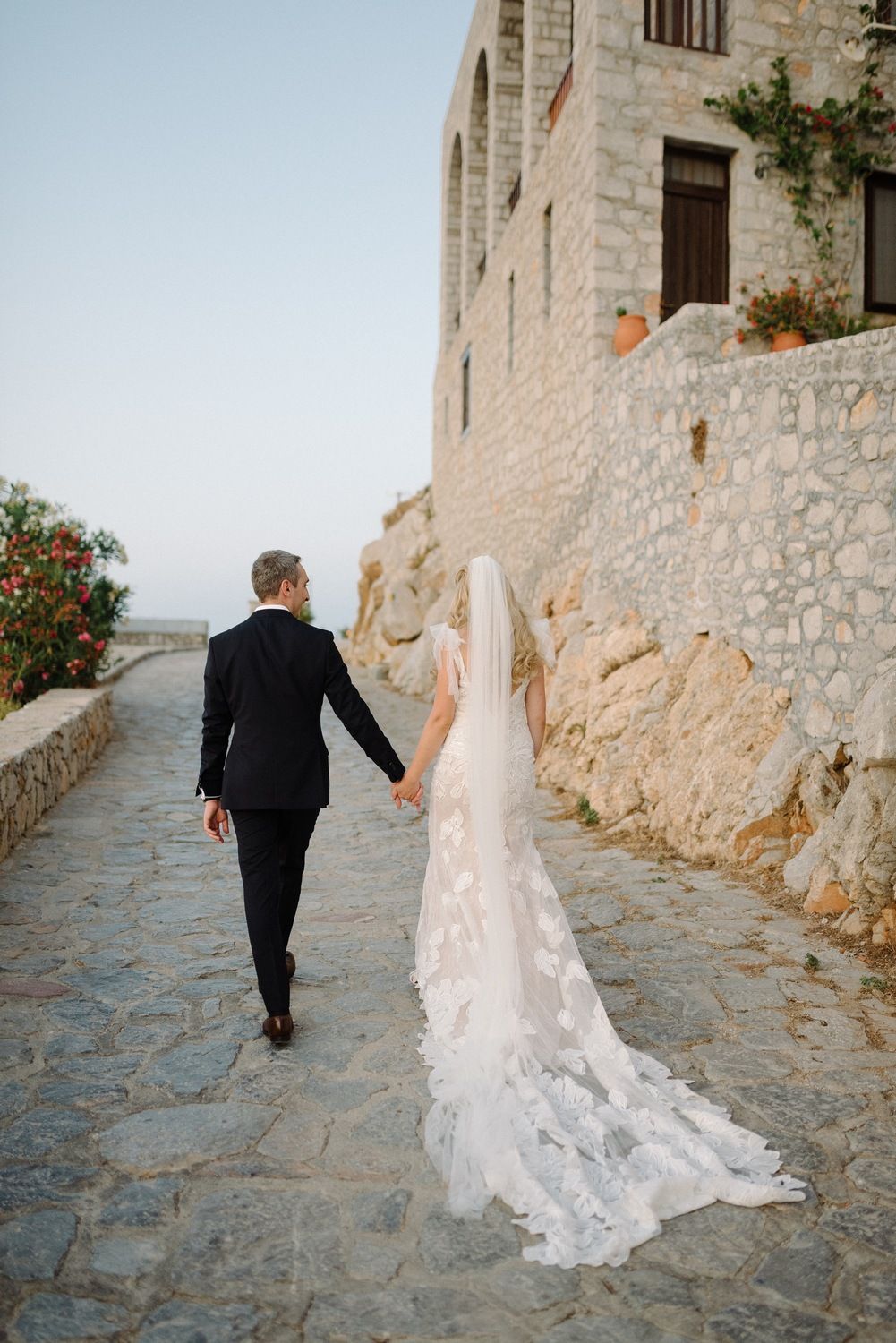 Wedding couple walks along stone pathway beside traditional Mediterranean stone building.