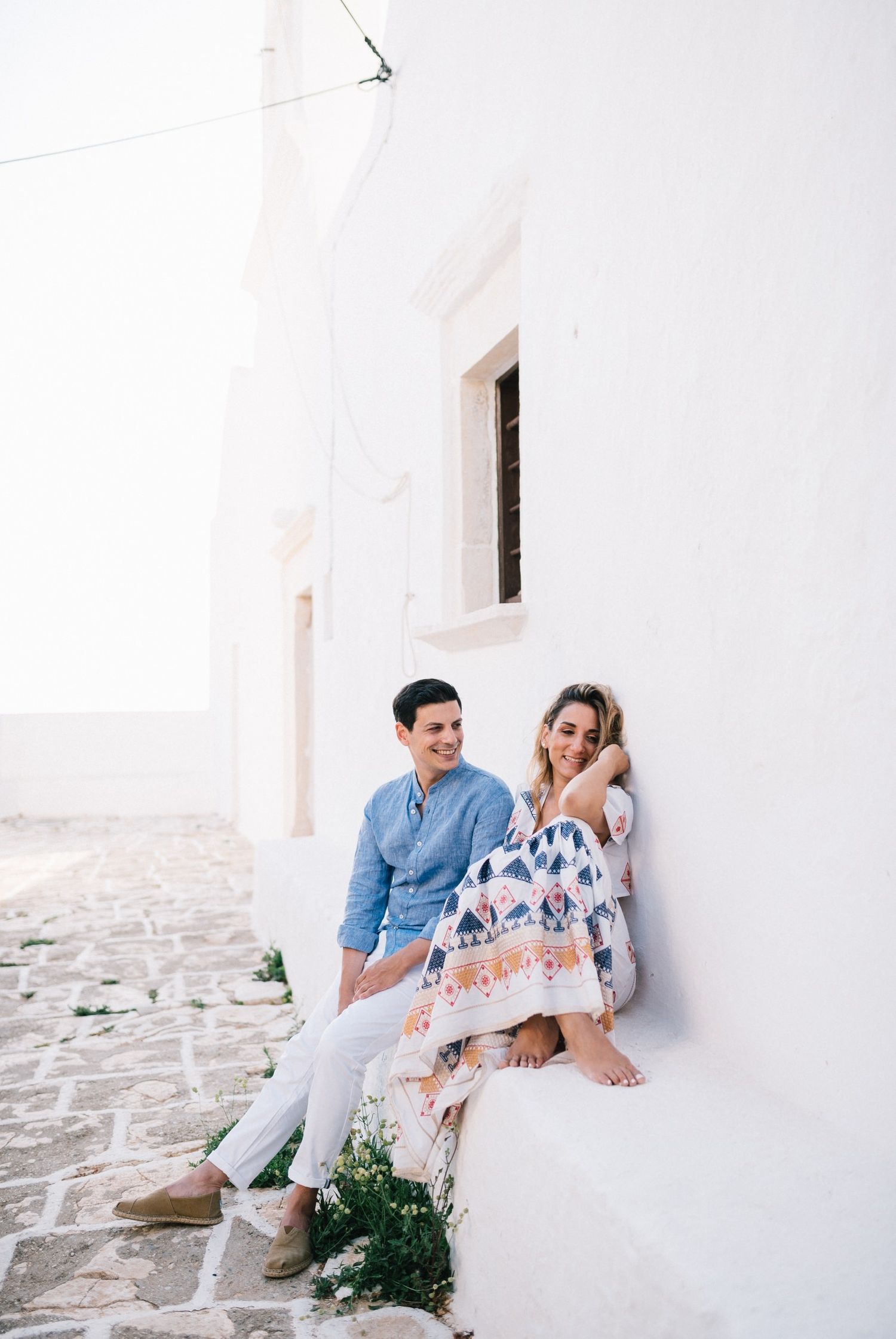 A relaxed couple sitting on a ledge against a white Mediterranean building wall.