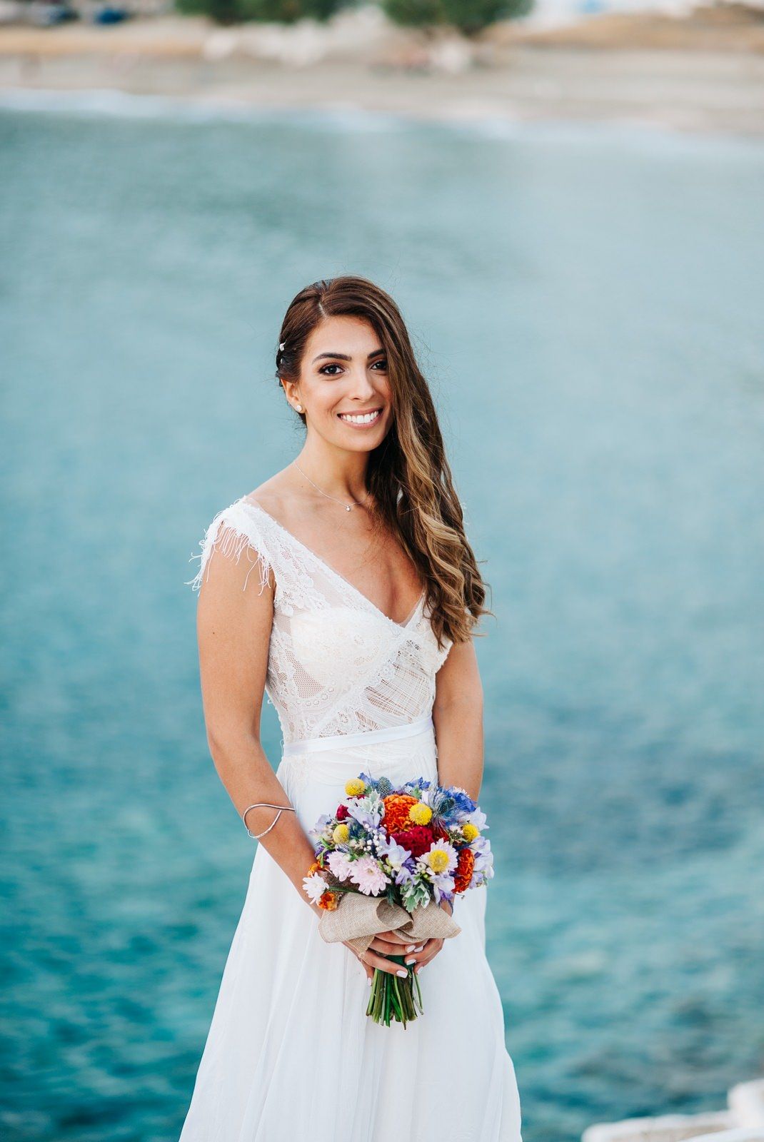 Beaming bride holds vibrant bouquet against stunning turquoise ocean backdrop.