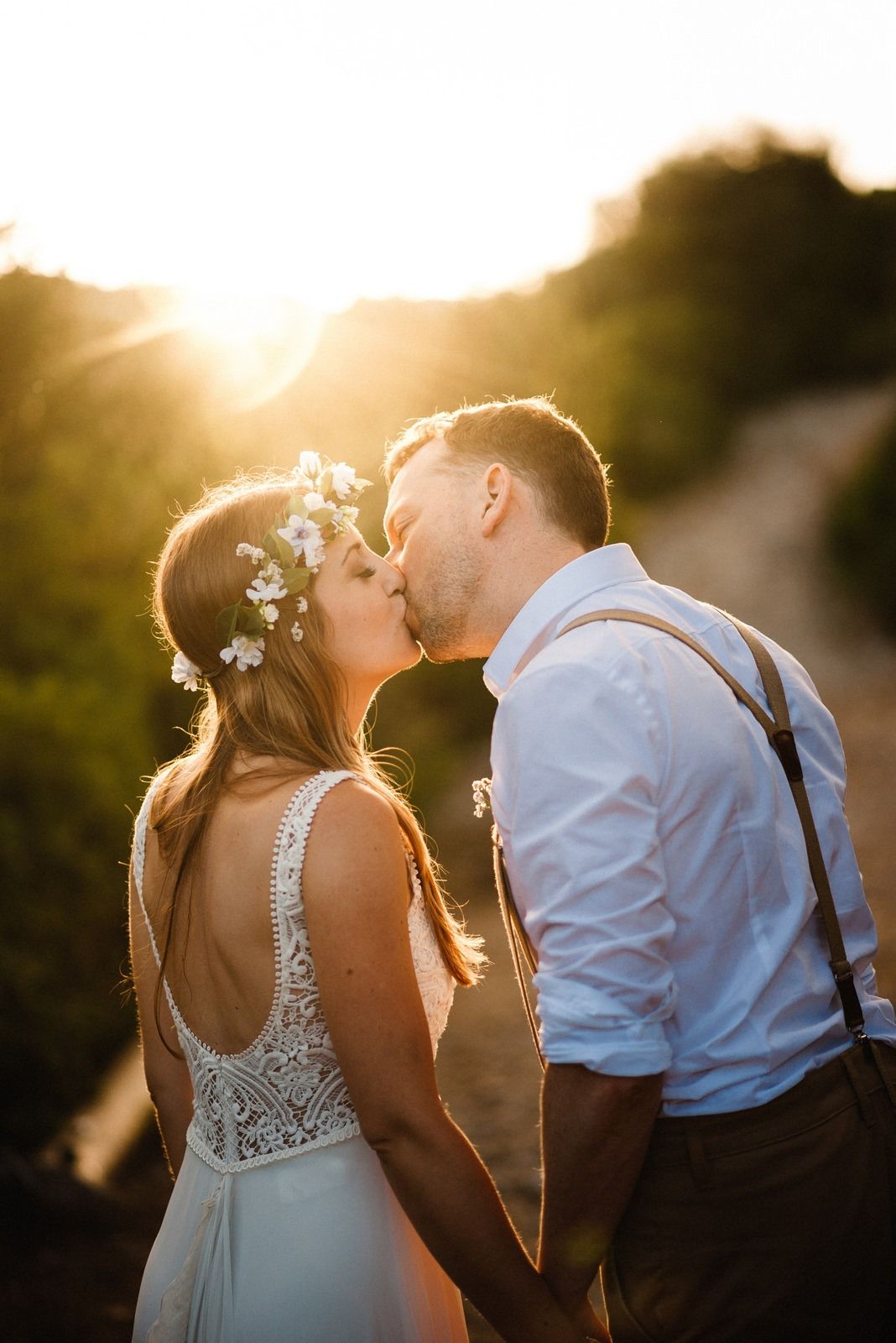Bride and groom share a romantic kiss backlit by golden sunlight on a nature path.