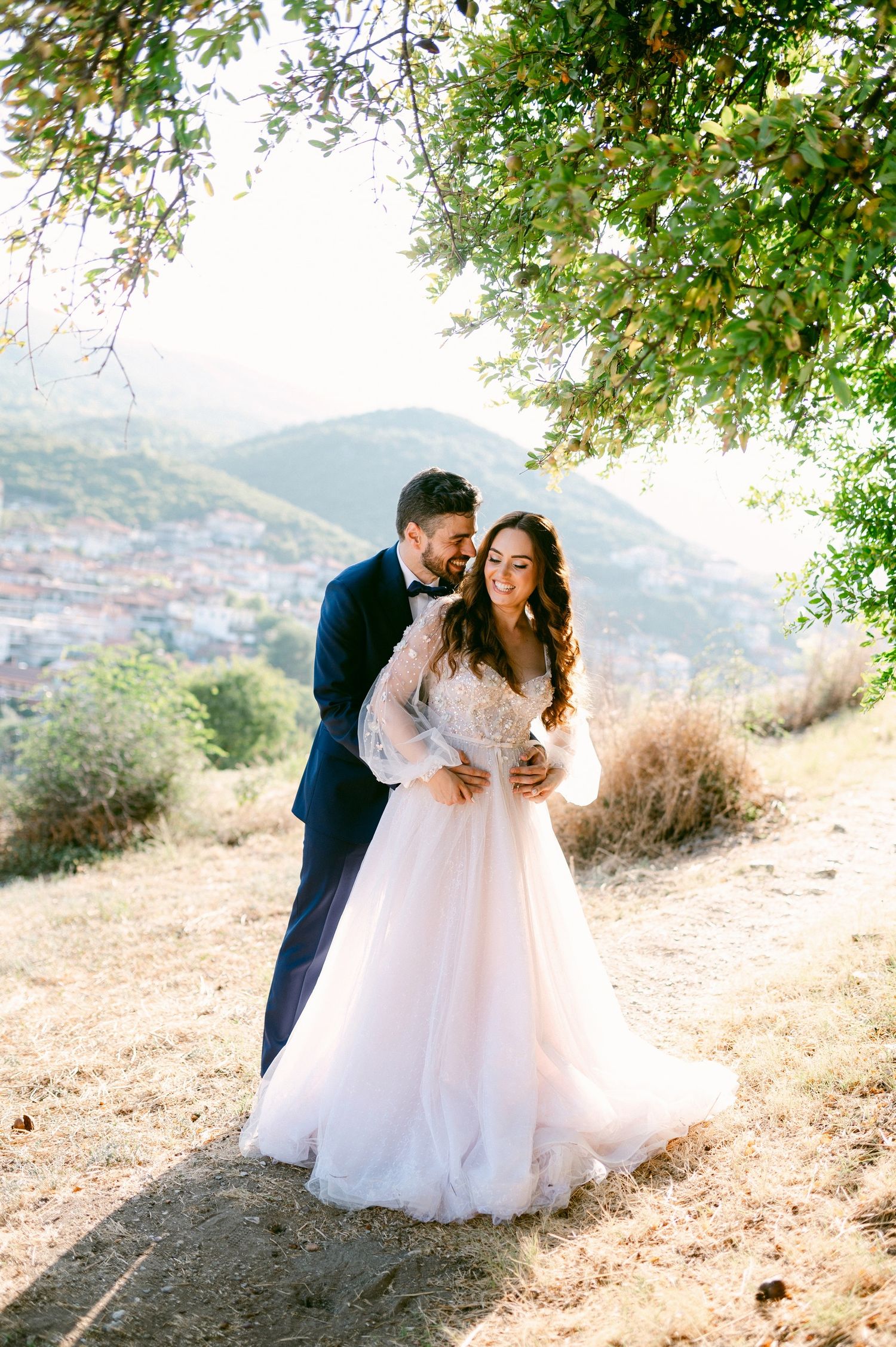 Couple in wedding attire share romantic moment on hillside overlooking Mediterranean town during golden sunlight.