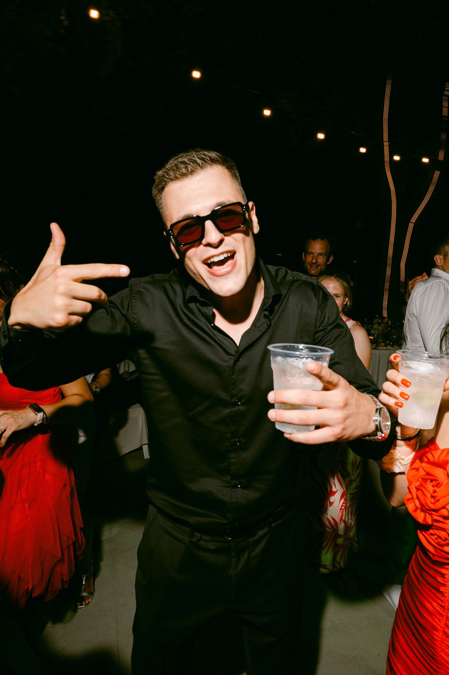 A party-goer in sunglasses and black attire gestures while holding a drink at a nighttime event.