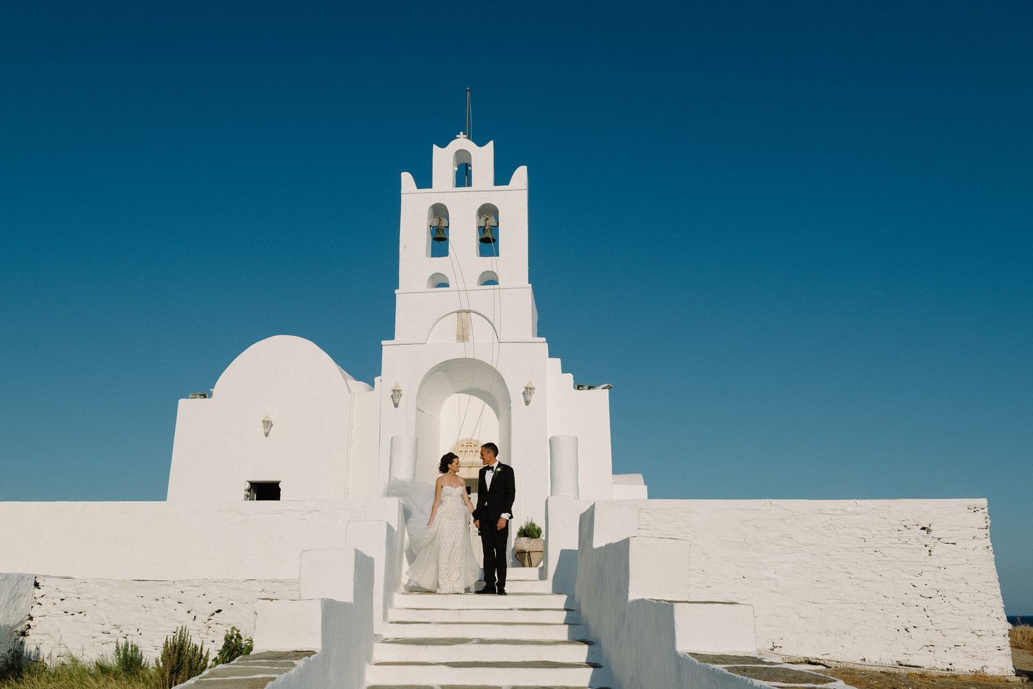 A couple stands at the entrance of Chrysopigi church in Sifnos with bell tower against bright blue sky in Greece.