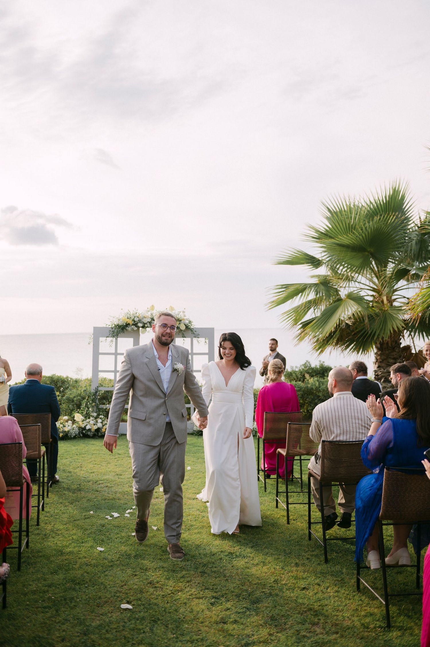A couple walks down the aisle at an outdoor wedding ceremony with palm trees and guests seated on both sides.