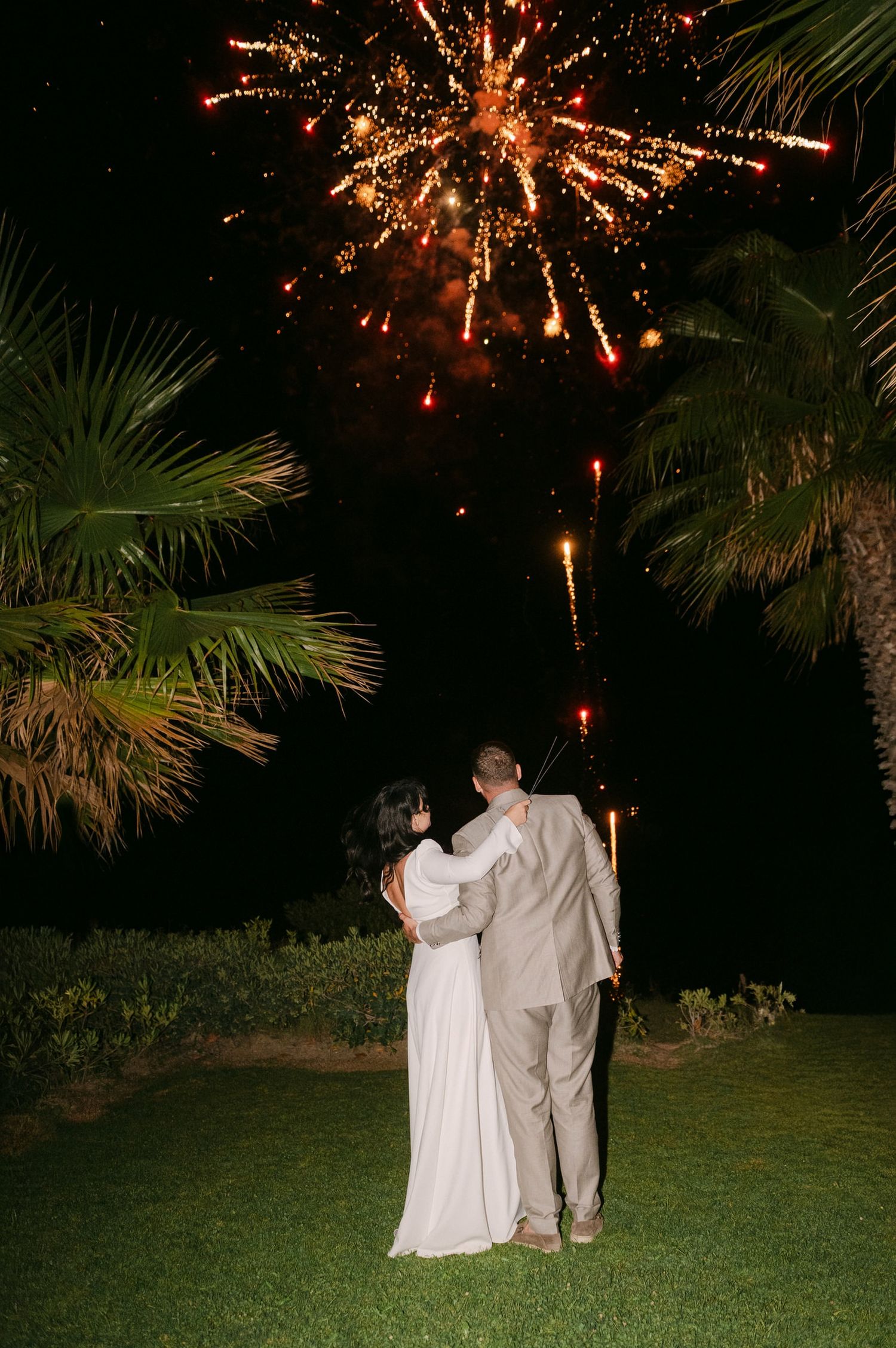 A couple in wedding attire watches fireworks burst in the night sky above palm trees during an outdoor celebration.