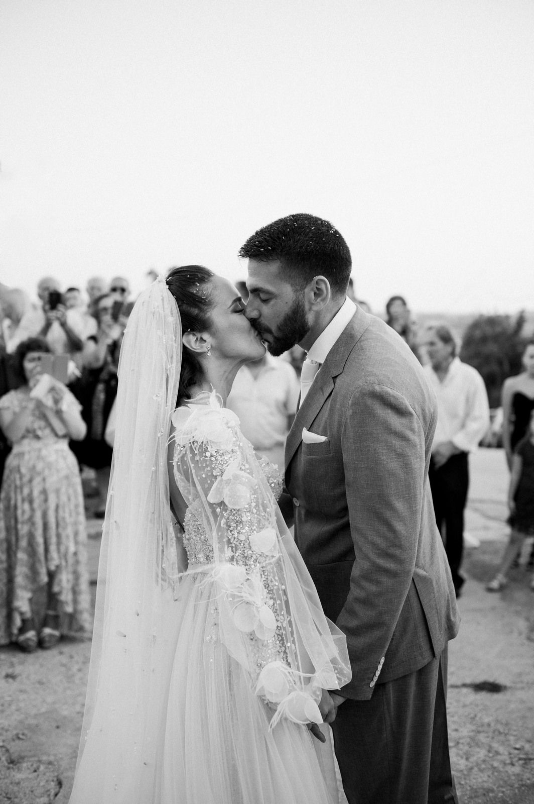 A romantic black and white wedding photo shows a couple sharing their first kiss as guests look on in the background.