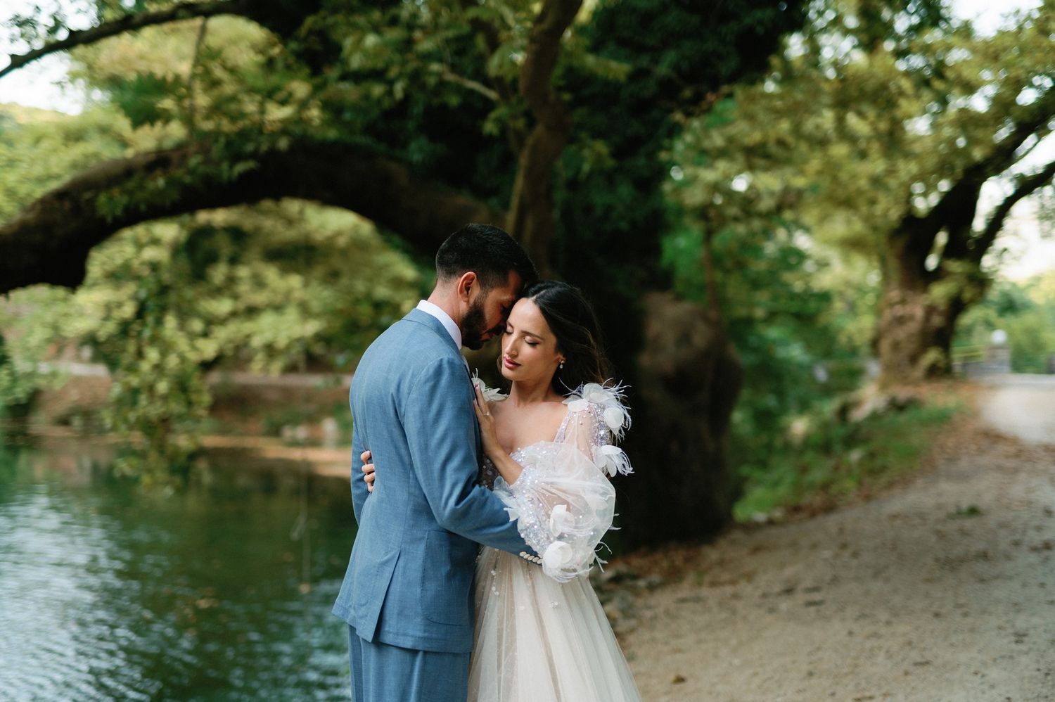 A romantic wedding couple shares an intimate moment by a tranquil lake surrounded by lush trees.