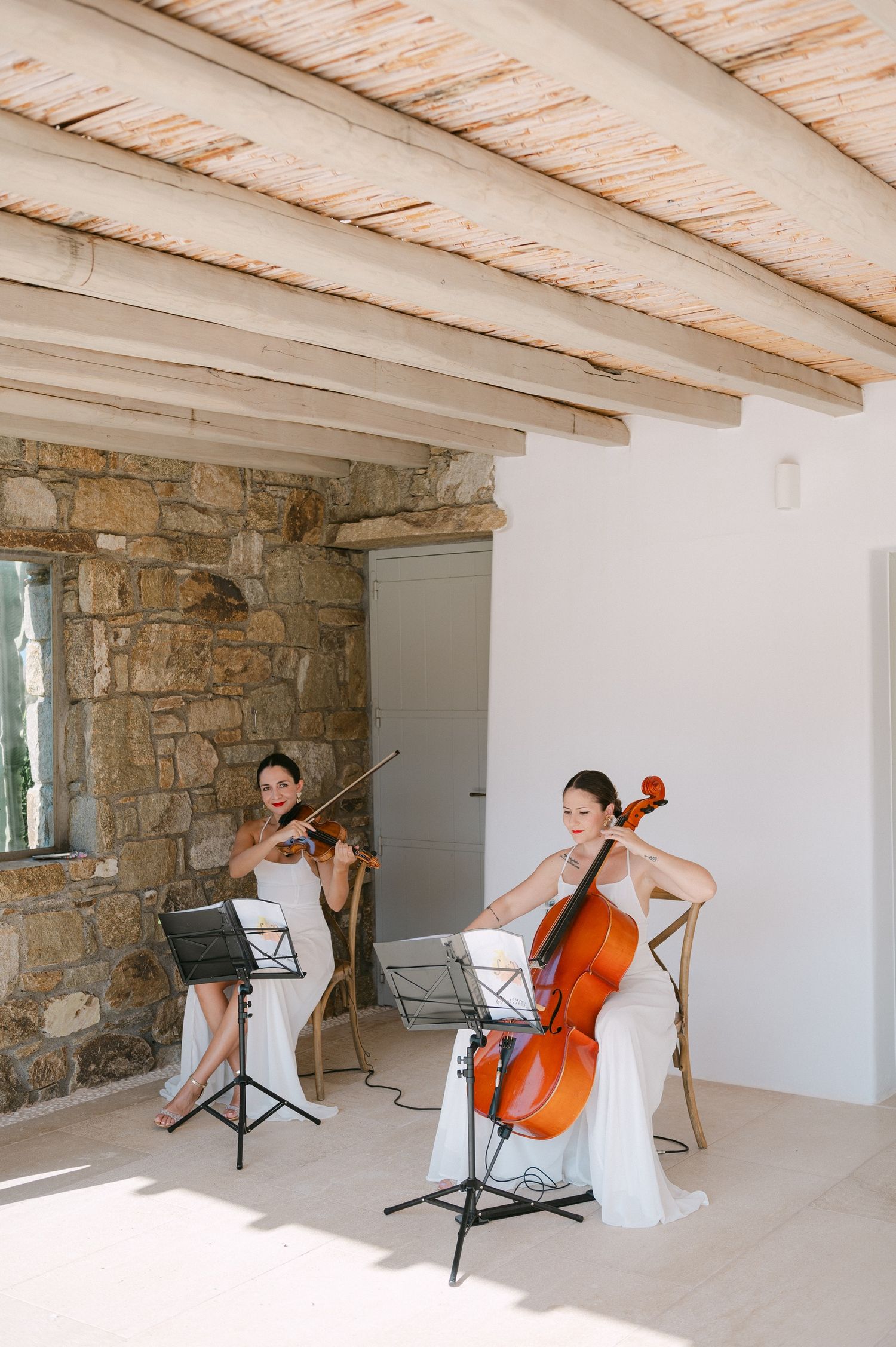 Musicians perform with string instruments in a rustic room with stone walls and wooden beamed ceiling.