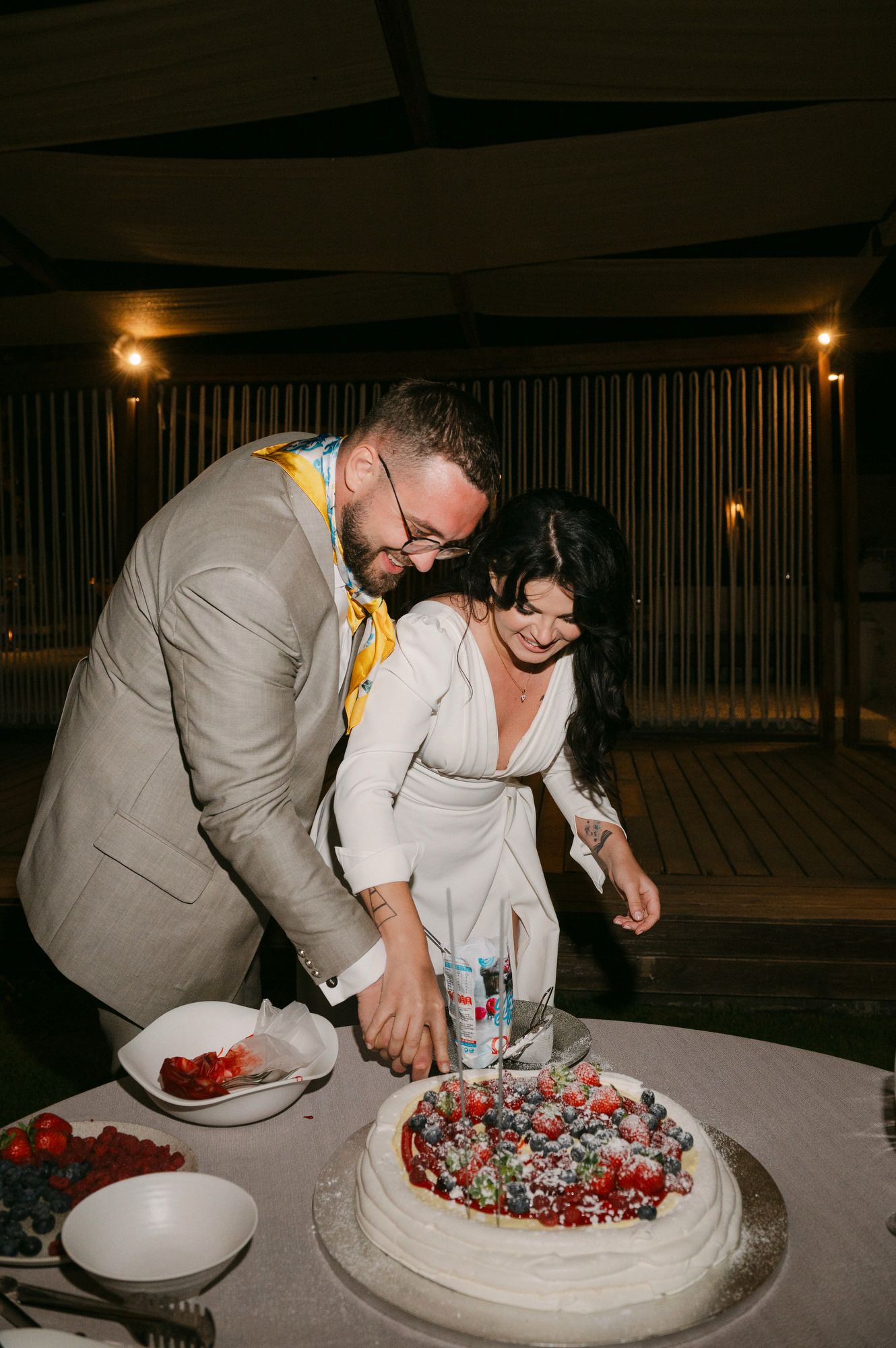 A couple cuts their wedding cake decorated with berries and cream at a dimly lit reception venue.