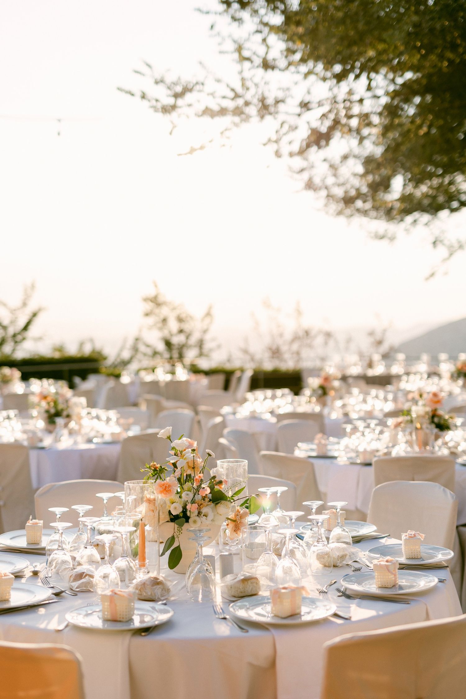 An elegant outdoor wedding reception setup with round tables decorated with floral centerpieces and white linens.