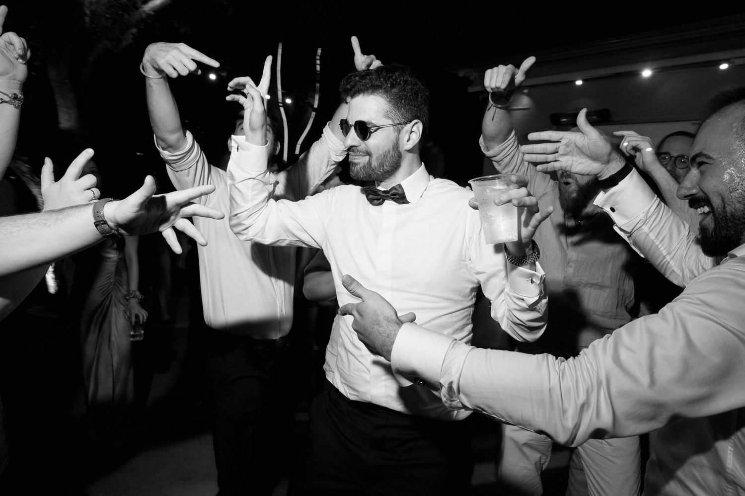 A lively black and white photo of a dancing crowd at a party with people raising their arms in celebration.