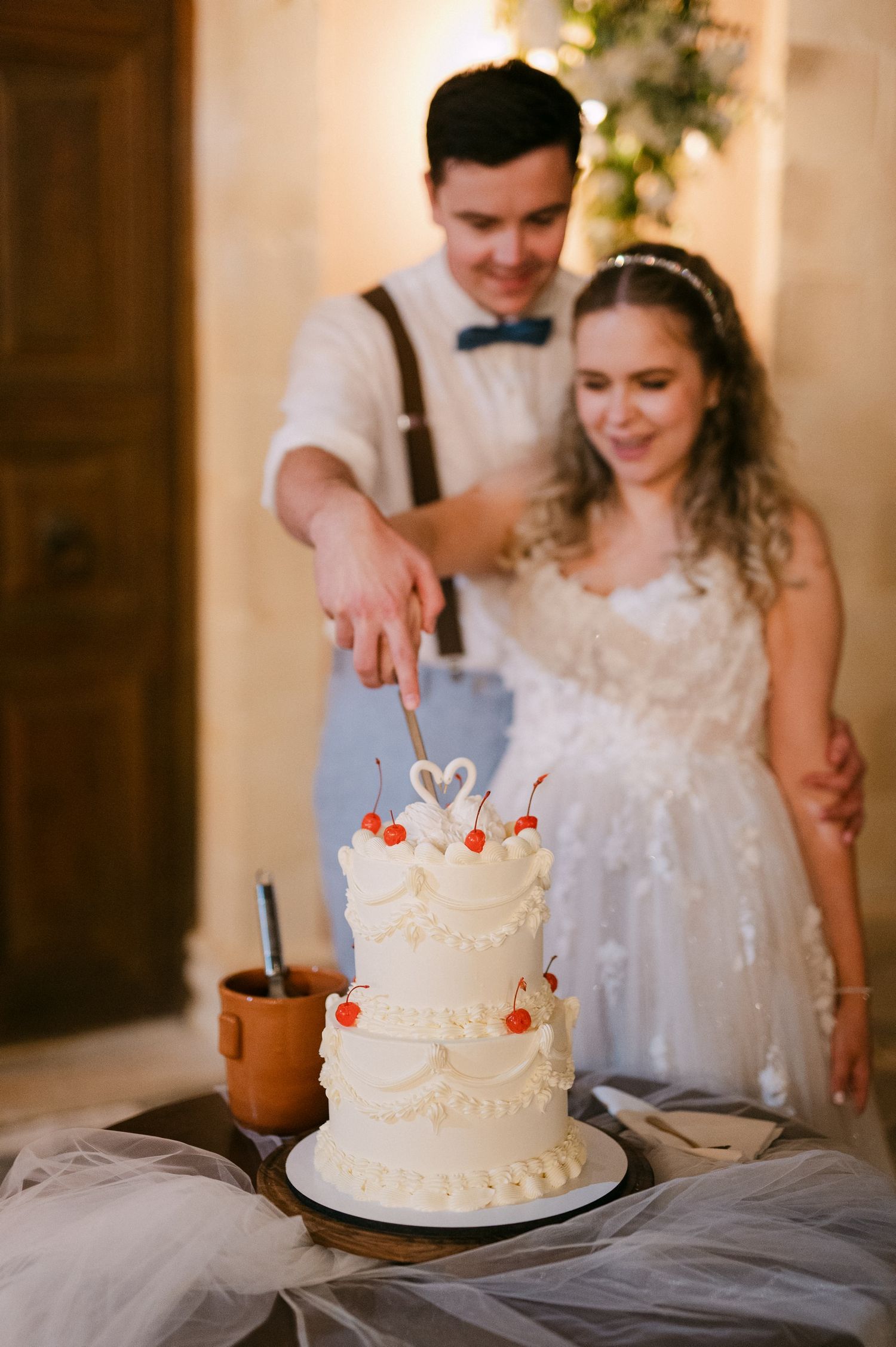 A couple cutting into their elegant white wedding cake decorated with swans and cherries on top during their reception.