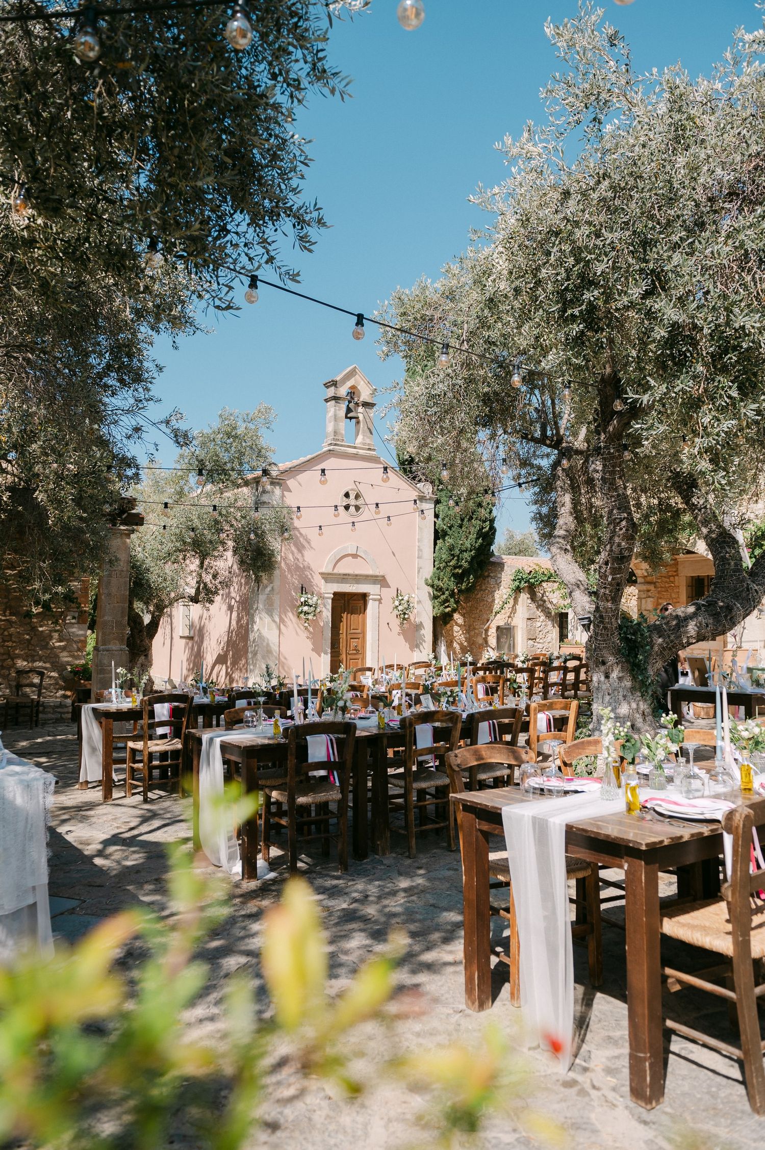 Multiple views of a rustic outdoor wedding reception with wooden tables under olive trees near a pink Mediterranean chapel.