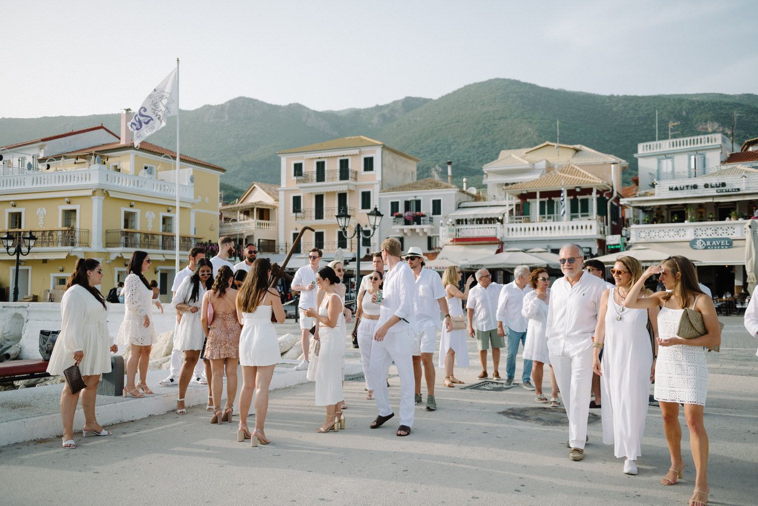 Group of people dressed in white gathering on a waterfront promenade with mountains in background.