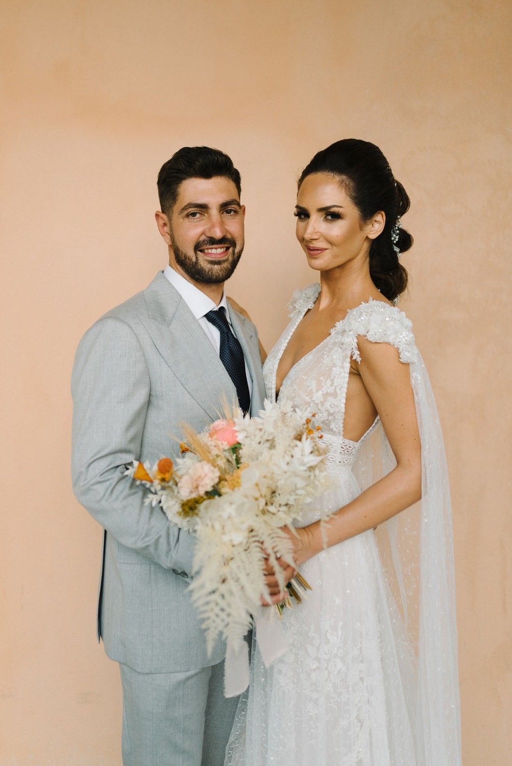 Elegant wedding couple holds a delicate bouquet featuring dried flowers, ferns and coral blooms against a peach wall.