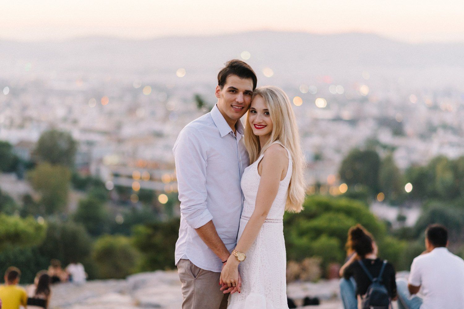 Couple posing together on hilltop with Athens cityscape and mountains in background.