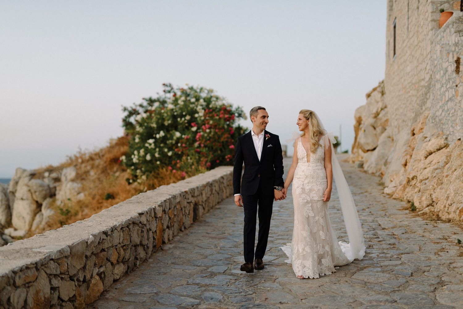 Newlyweds stroll along picturesque Mediterranean pathway with stone wall and flowering bushes.
