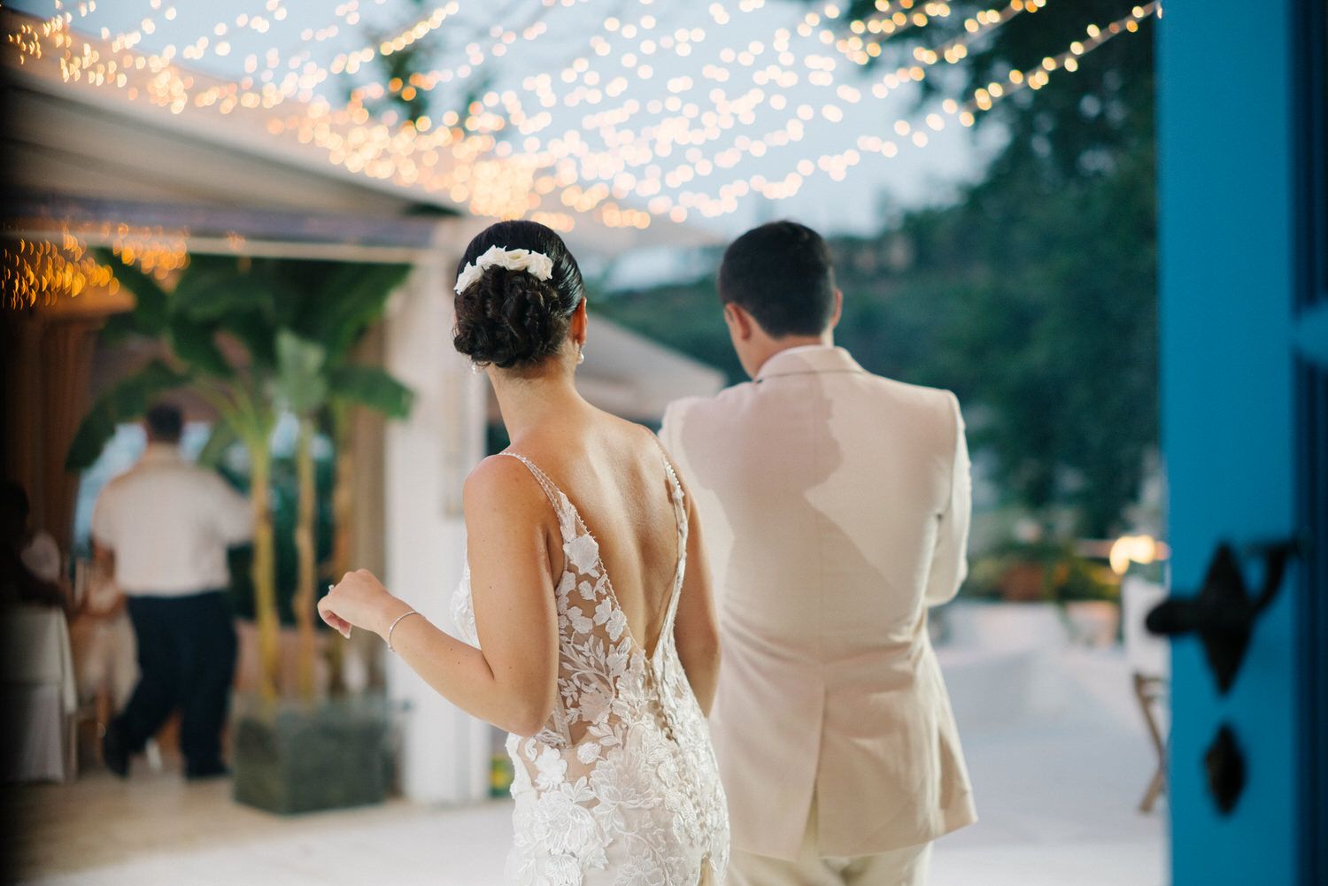 Elegant couple walks together under twinkling reception lights in formal wedding attire.