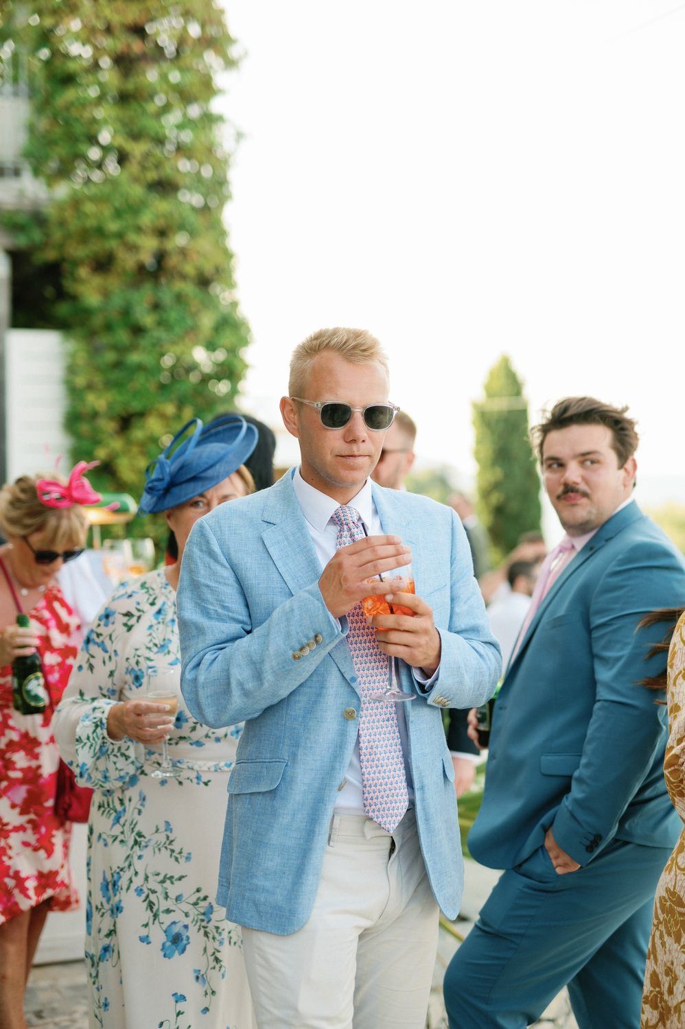 Guests enjoying cocktails at an elegant outdoor party with men in suits and women in colorful dresses.