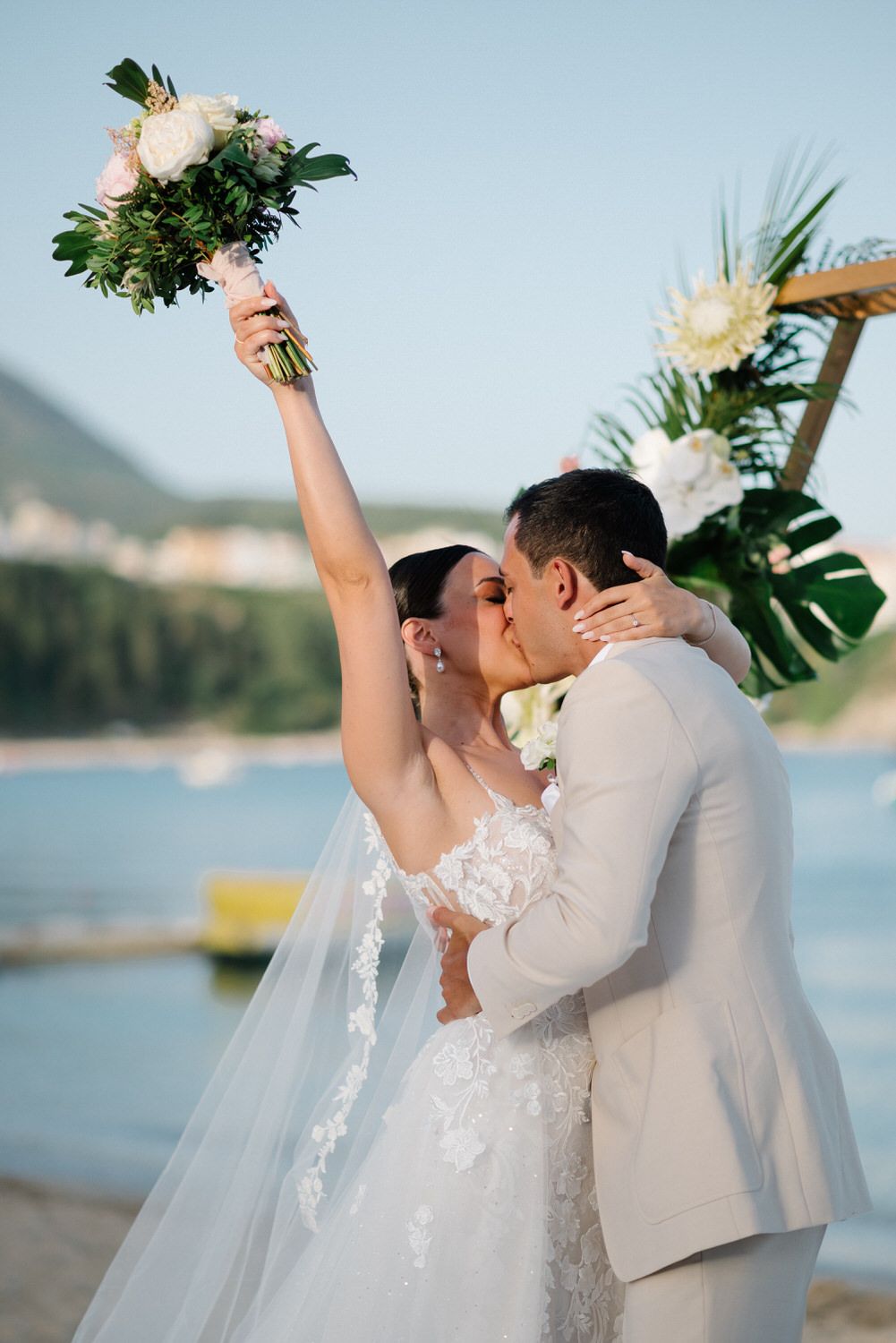 Newlyweds celebrate with a passionate kiss as bride raises her bouquet against ocean backdrop.