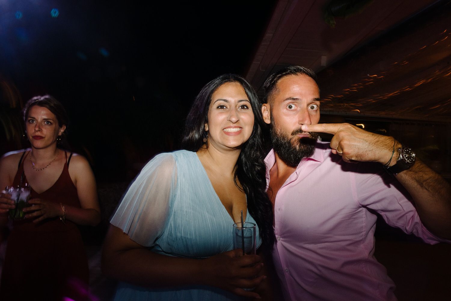 Party guests enjoy drinks and dancing at a lively indoor reception with colorful lighting.