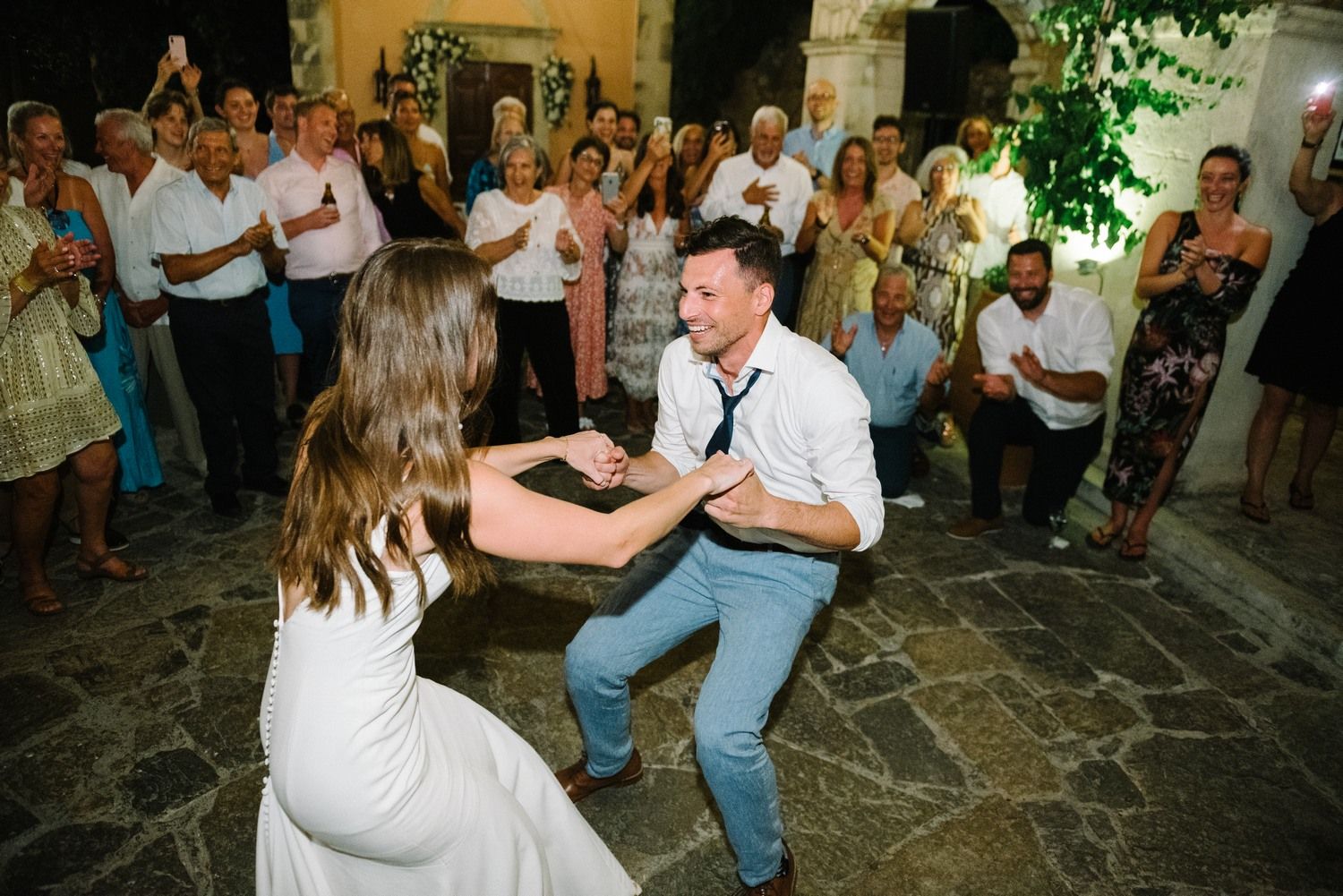 Couple performing energetic dance moves on stone patio while wedding guests cheer.