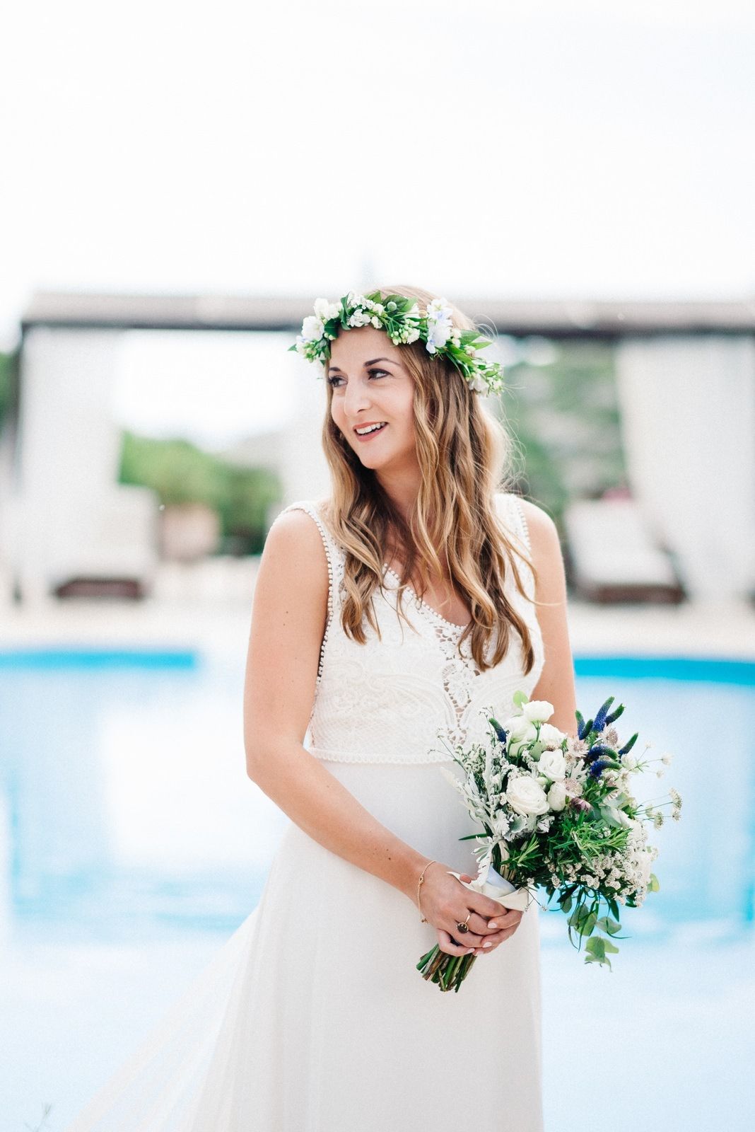Bride in lace gown and floral crown smiling by pool with white bouquet.