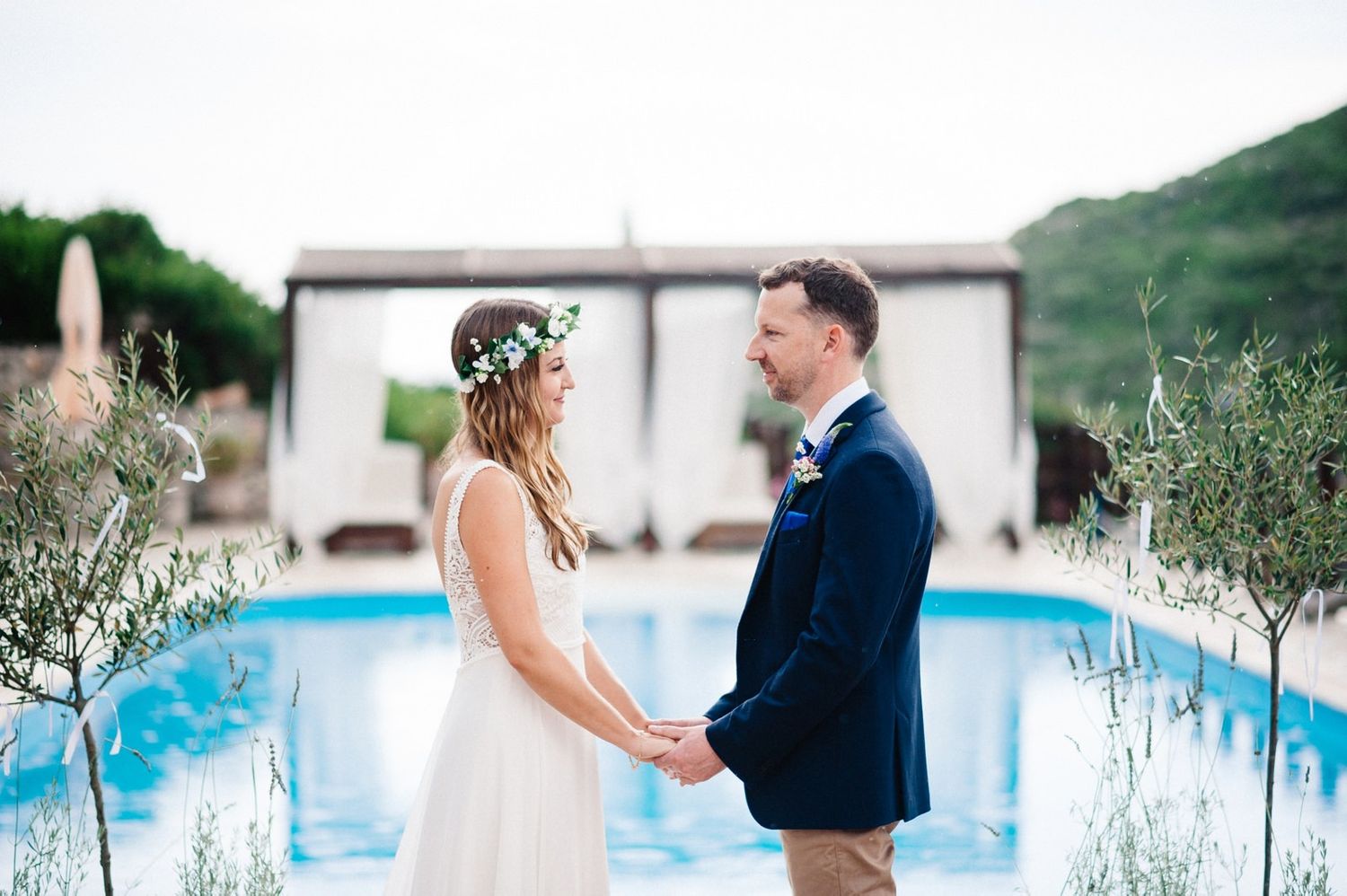Intimate wedding ceremony moment by pool with olive branches and mountain scenery.