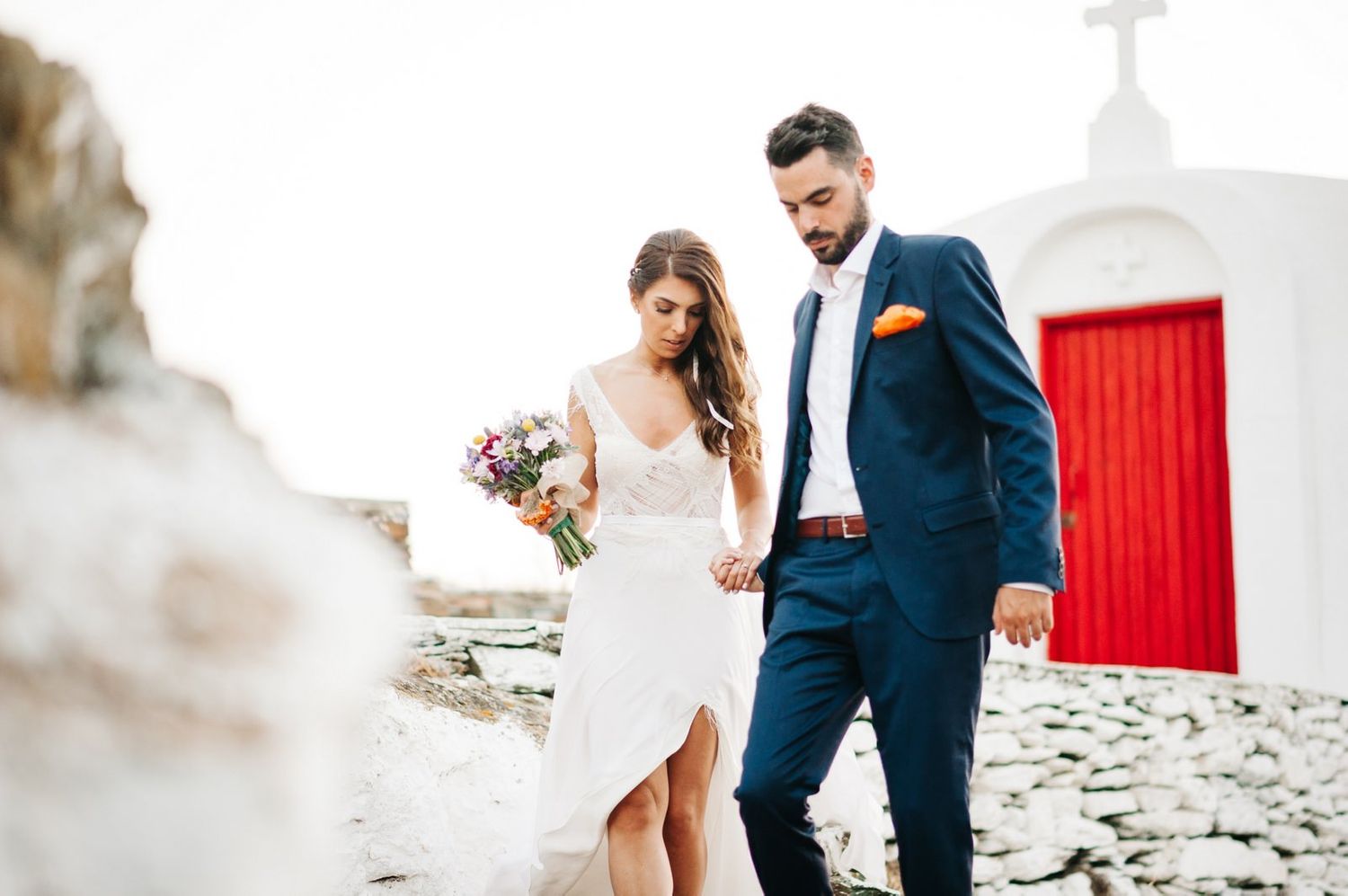 Newlyweds walking together on traditional Greek island pathway with red church door.