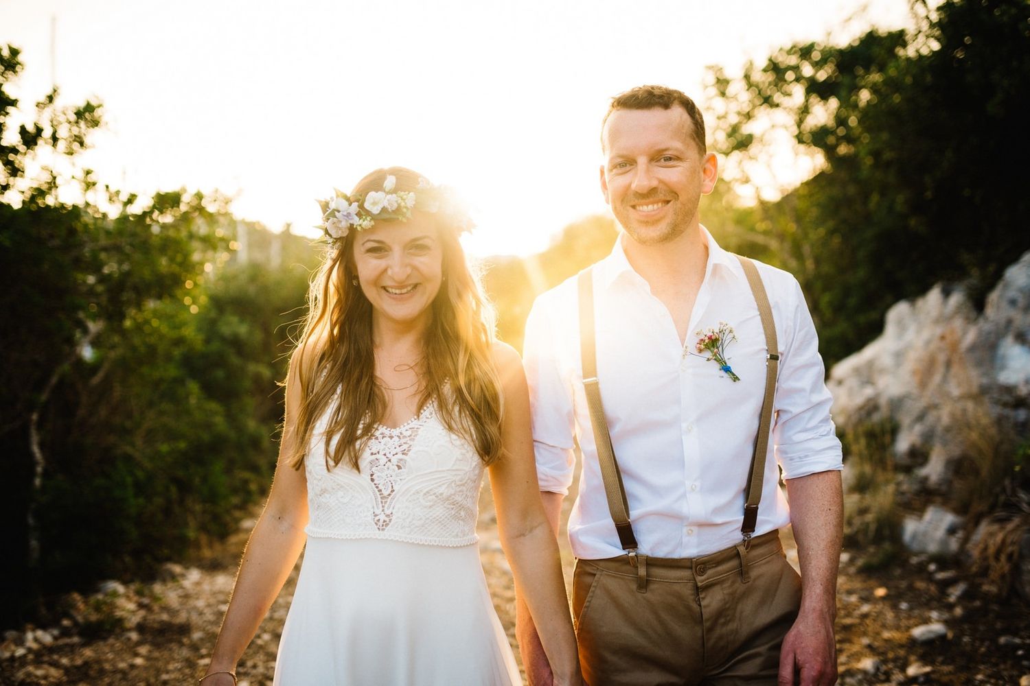 Happy couple in wedding attire smiling together on a sunny mountain path.