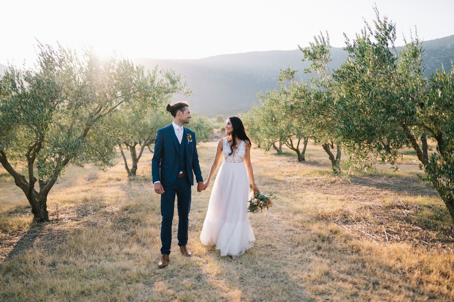 Bride and groom walk hand in hand through olive grove during sunset.
