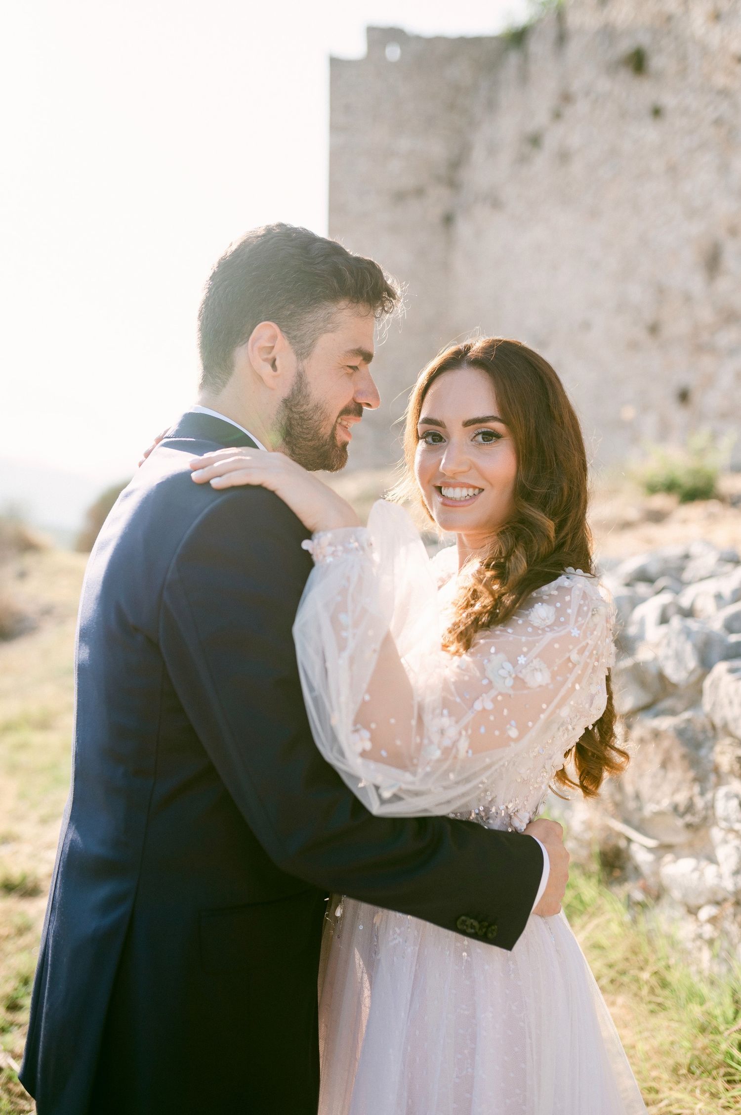 A romantic wedding photo of a couple embracing outdoors with sunlight and an old stone wall in the background.