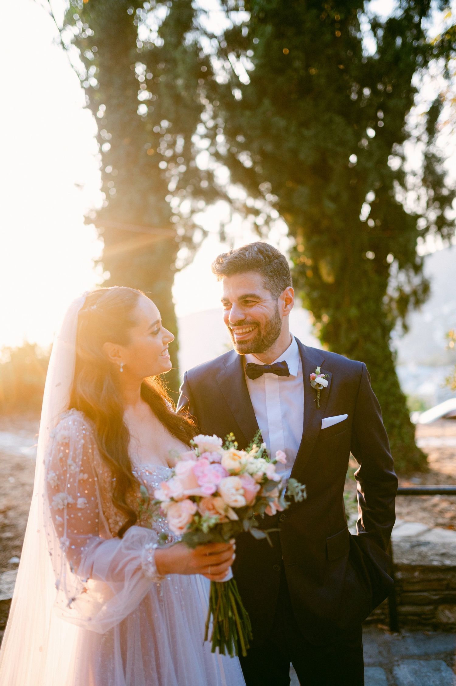 A happy newlywed couple shares a candid moment in the golden sunlight with the bride holding a pink bouquet.