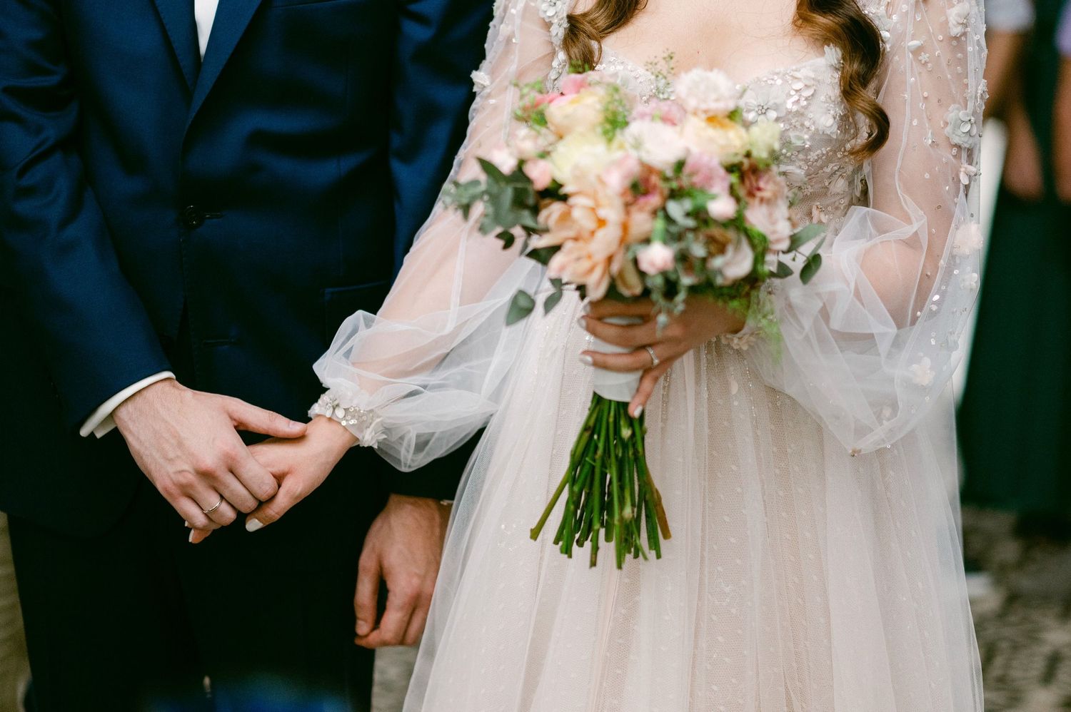 Close-up view of wedding couple holding hands with elegant white bouquet and lace sleeve details.