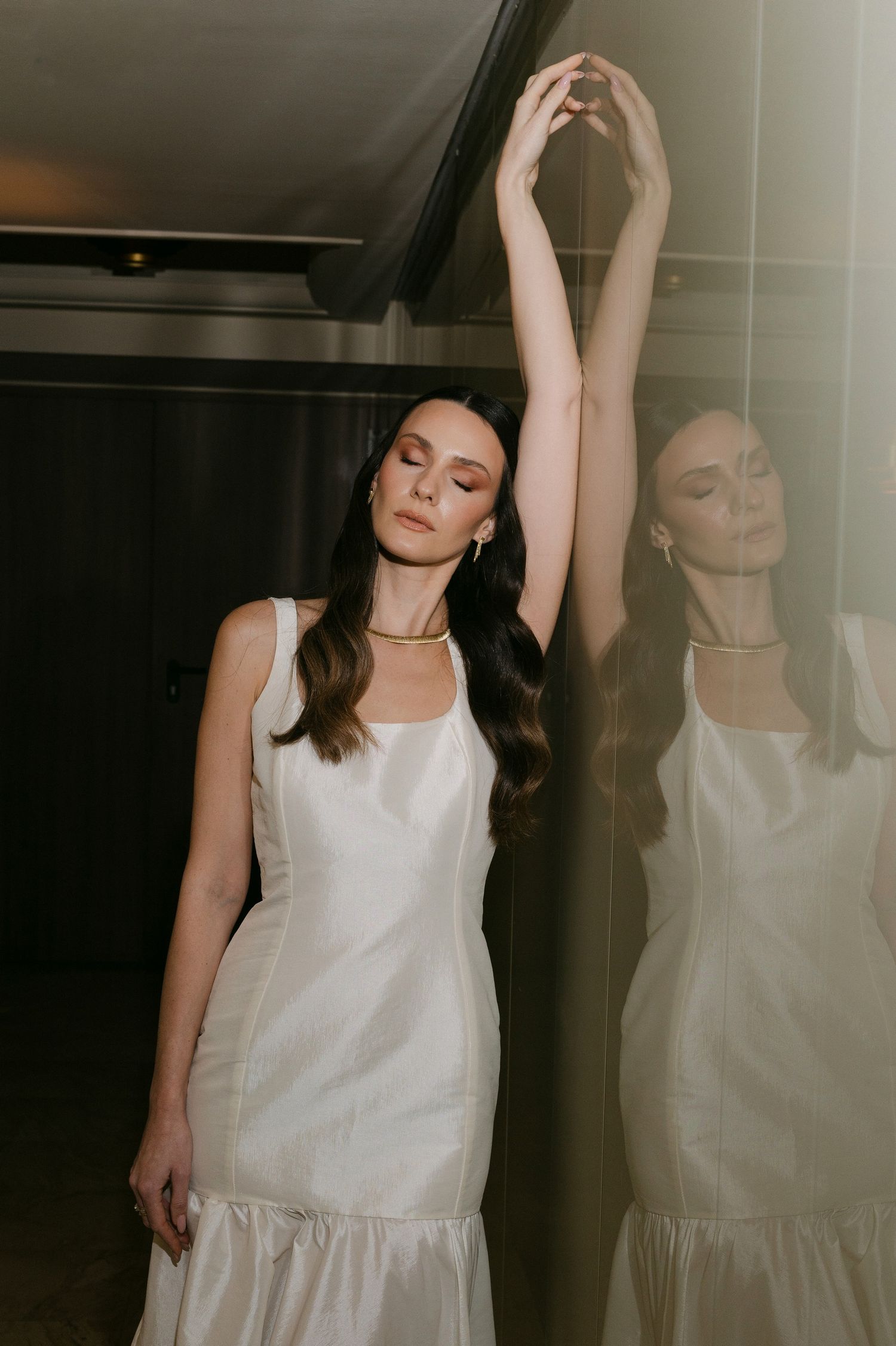 A woman in a white sleeveless dress poses gracefully against a wall with her reflection visible in dramatic lighting.