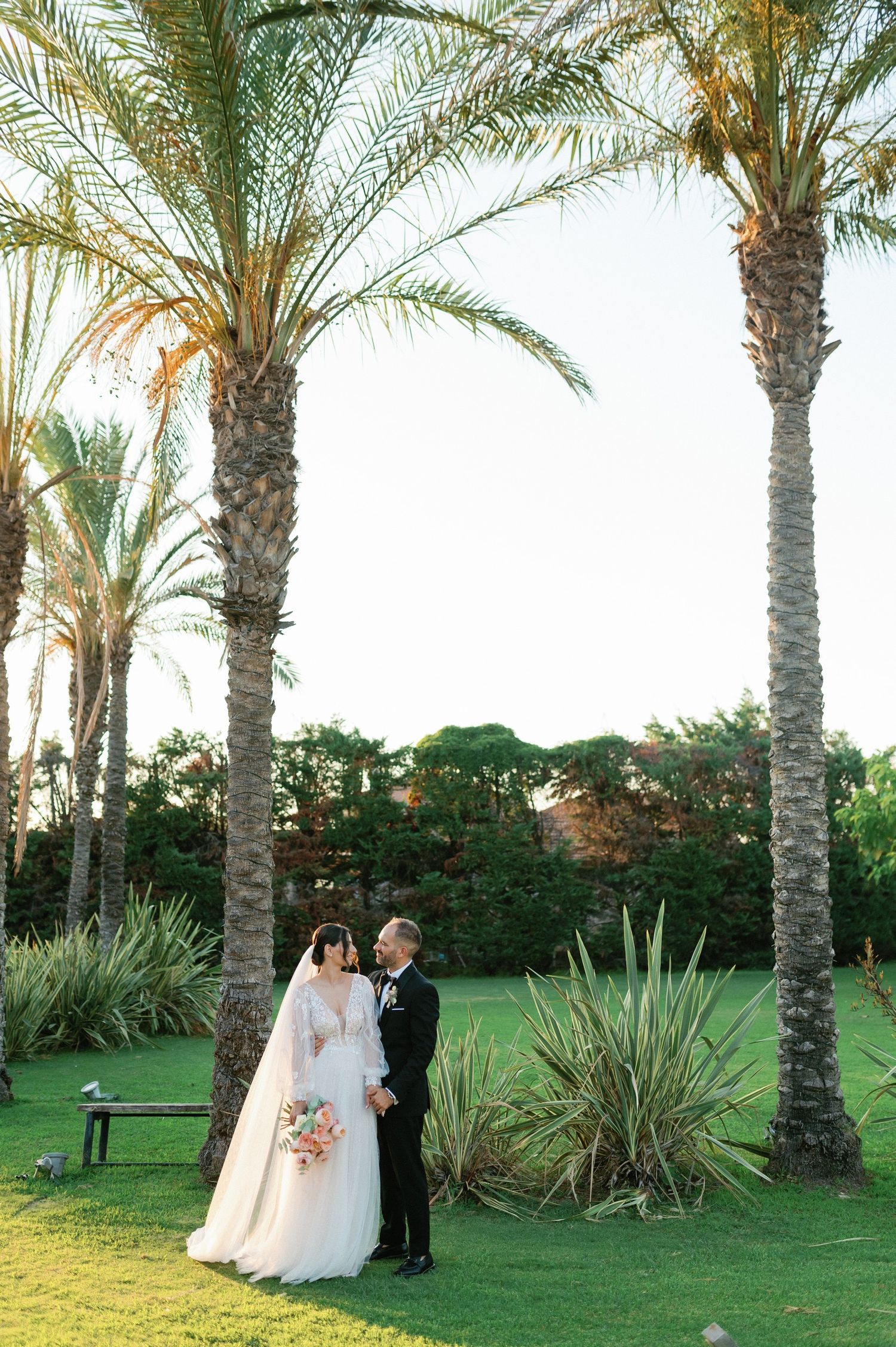 A romantic wedding couple shares a tender moment among tall palm trees in a lush garden setting.