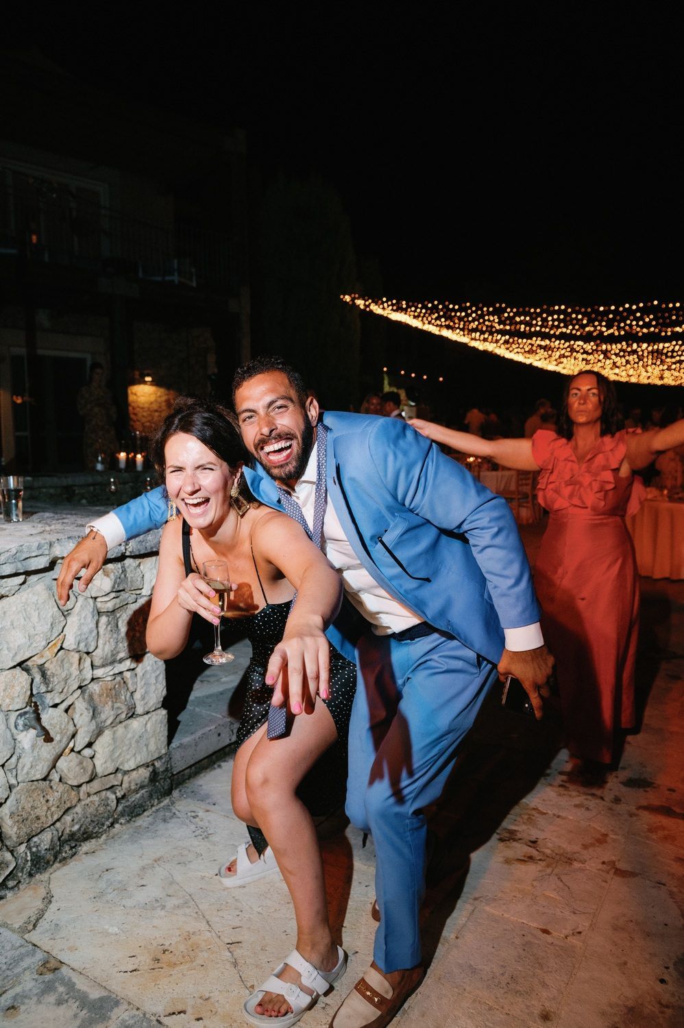 Party guests pose and dance along a stone wall at a nighttime celebration.